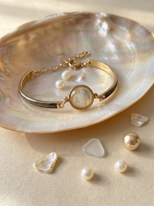 A gold-toned  plated and rhodium plated bangle bracelet with a round iridescent nautilus centerpiece, displayed on a large iridescent shell with small pearls scattered around on a warm beige surface.