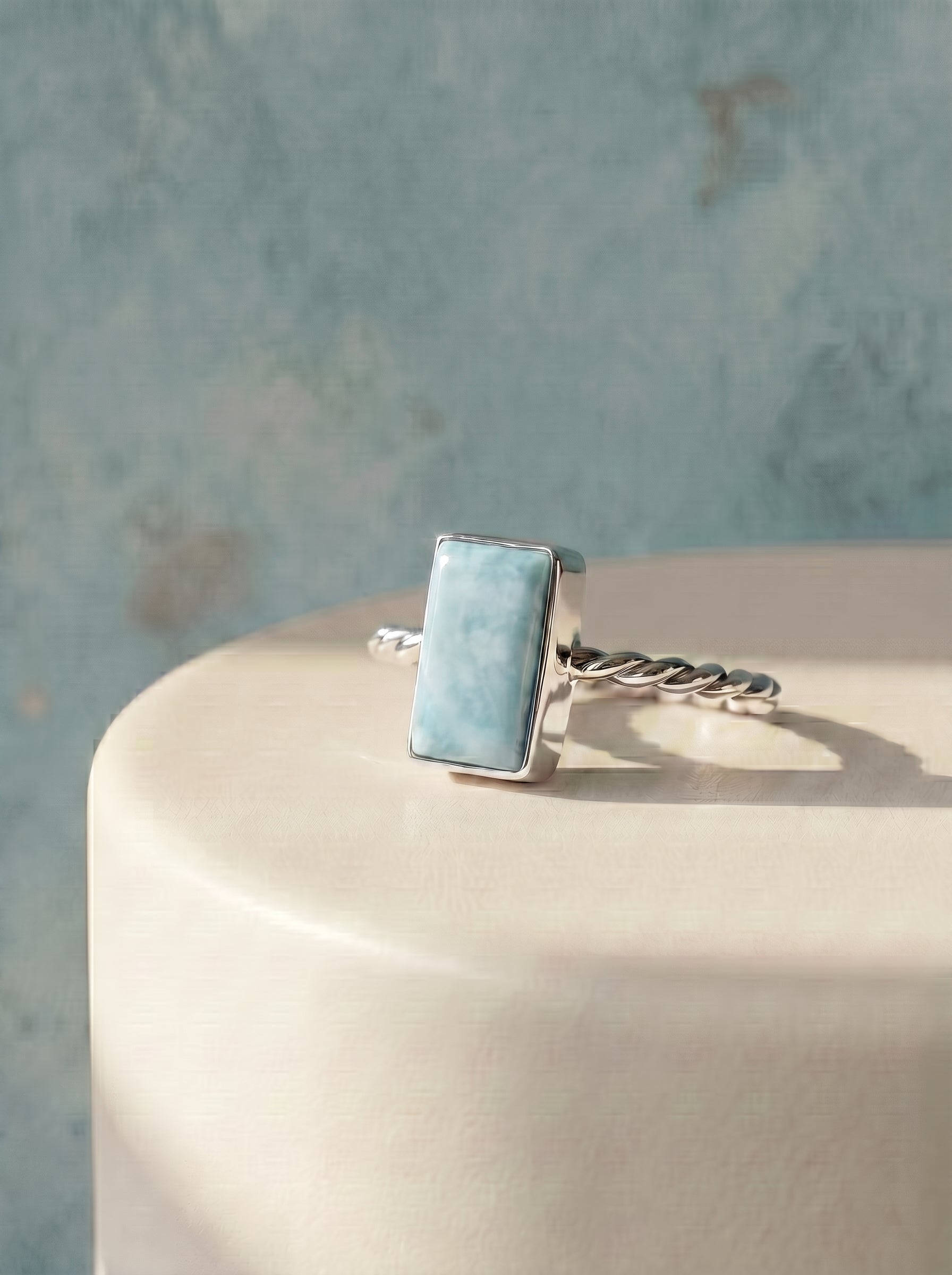 A silver ring with a rectangular light blue Larimar stone, displayed on a cream-colored pedestal against a textured blue background.