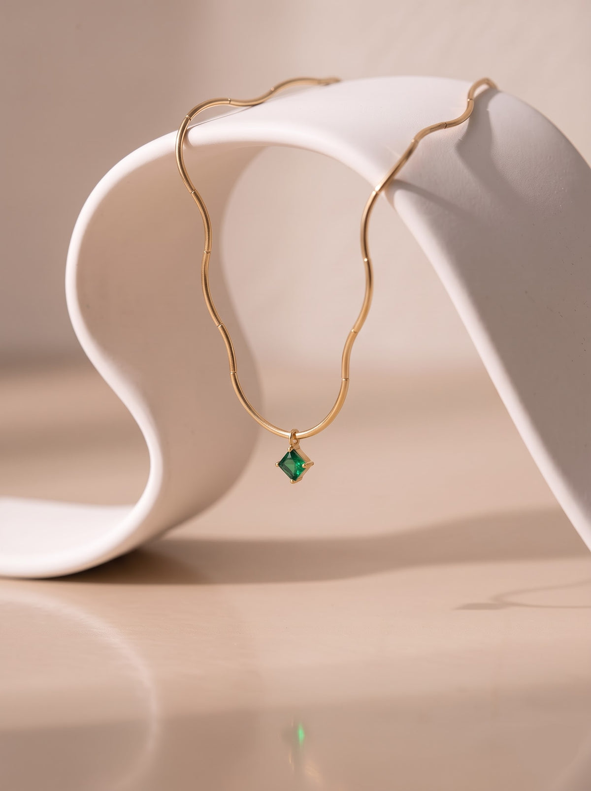 StoneCraft Jewellery gold wave chain necklace with a square green crystal pendant displayed on a white dish beside small flowers.