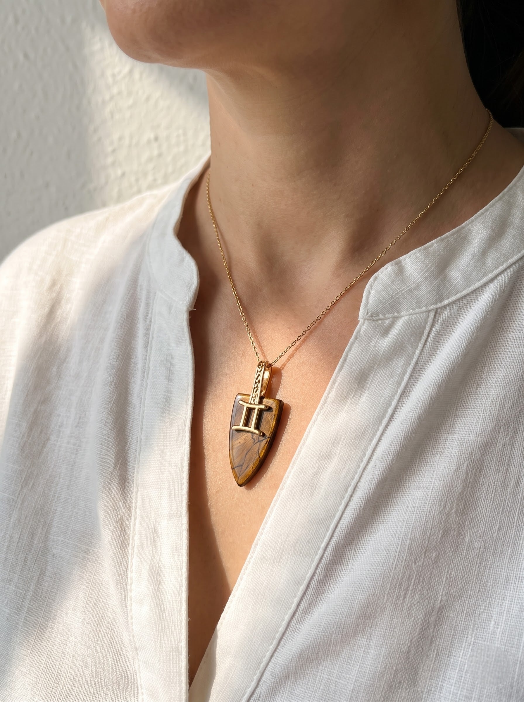 Woman wearing StoneCraft Jewellery tiger's eye Gemini zodiac shield pendant necklace on gold chain, white linen top close-up.