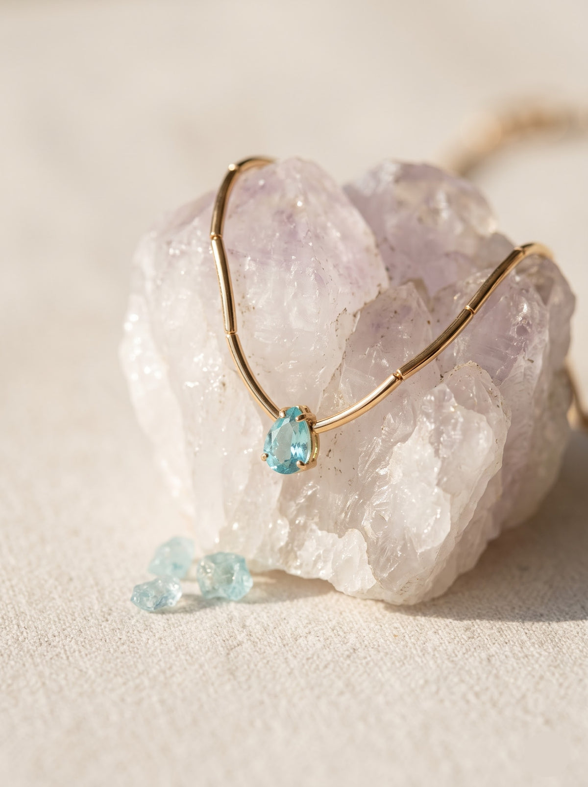 StoneCraft Jewellery gold wave chain necklace with a light blue teardrop crystal pendant displayed on an upright white stand.