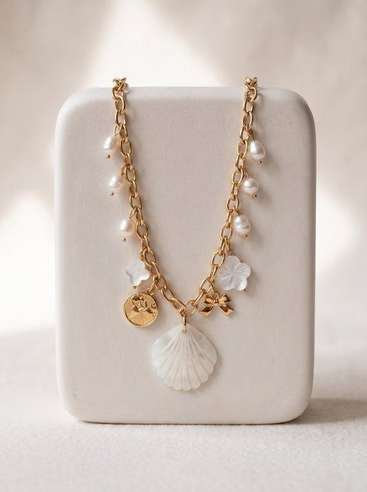 Gold-plated charm necklace with mother of pearl shell pendant, freshwater pearl and flower charms displayed on a white jewellery stand