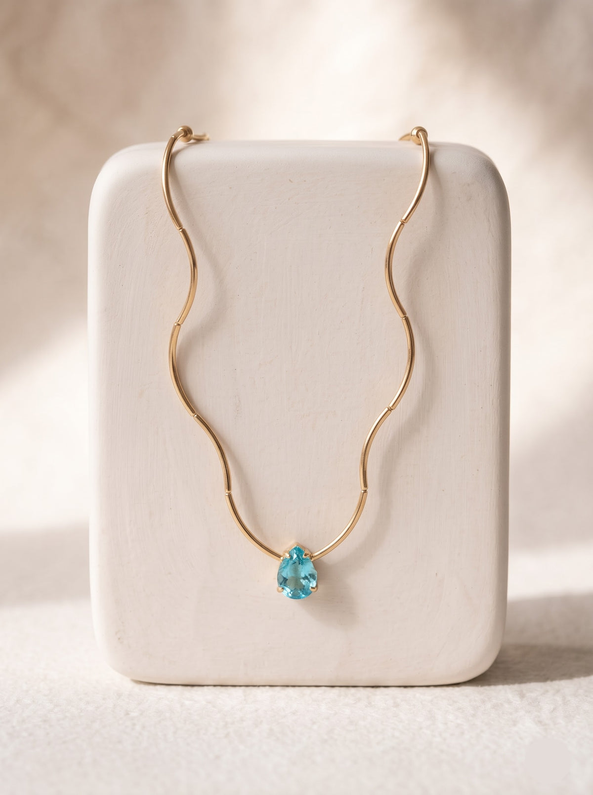 Woman wearing a StoneCraft Jewellery gold wave chain necklace with a light blue teardrop crystal pendant and a white halter neckline.