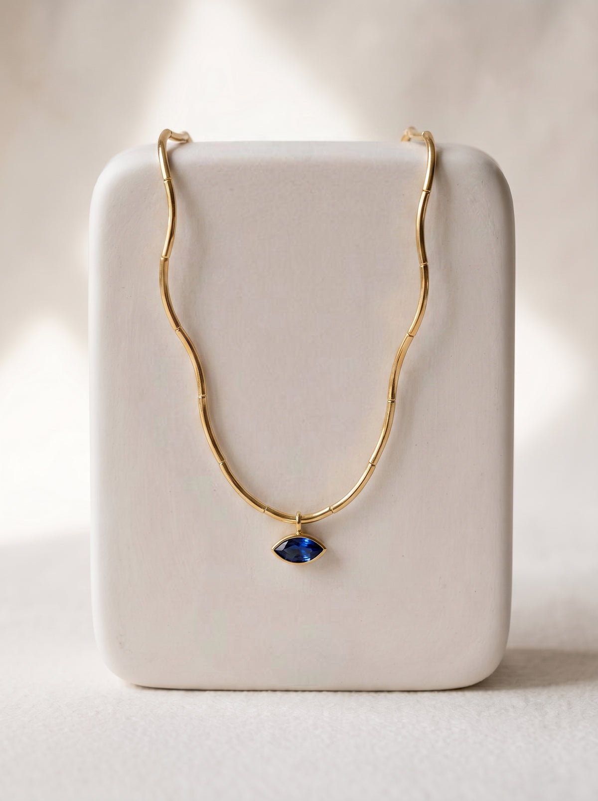 A delicate gold wavy chain necklace with a small blue teardrop-shaped pendant, displayed on a white rectangular jewelry stand against a softly lit neutral background.