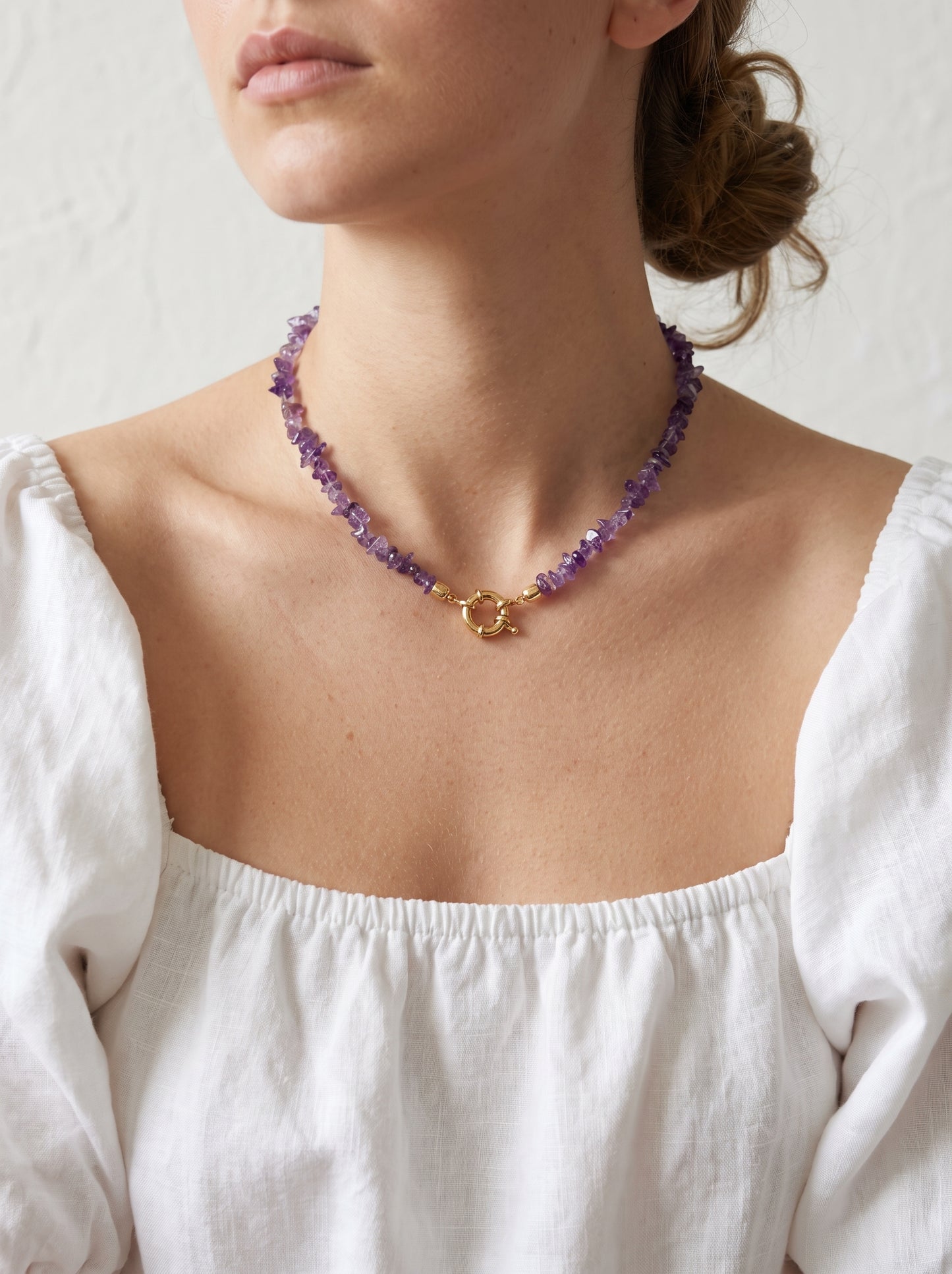A close-up of a model wearing a delicate necklace made of purple amethyst beads with a gold clasp, paired with a white off-the-shoulder blouse.