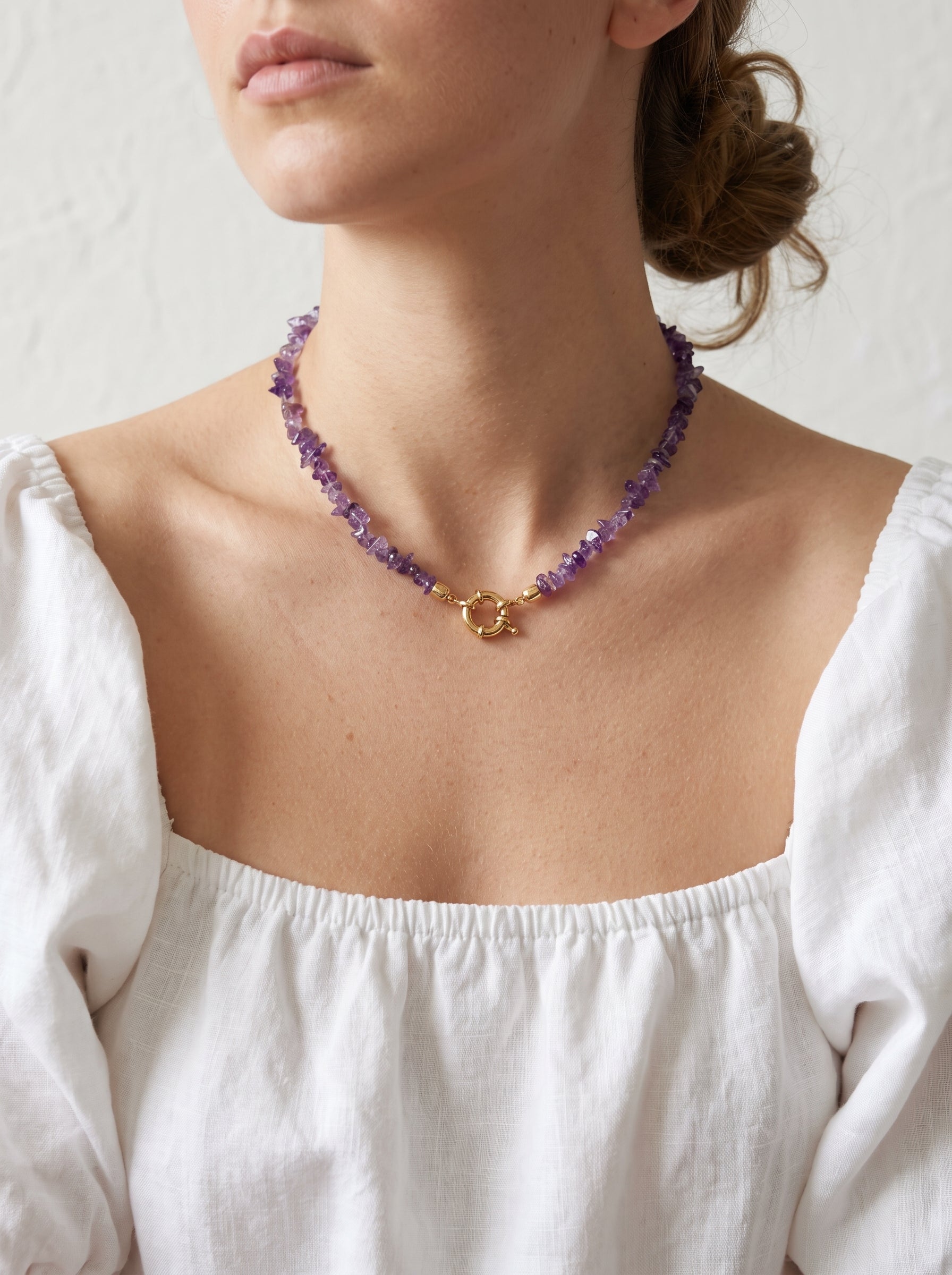 A close-up of a model wearing a delicate necklace made of purple amethyst beads with a gold clasp, paired with a white off-the-shoulder blouse.