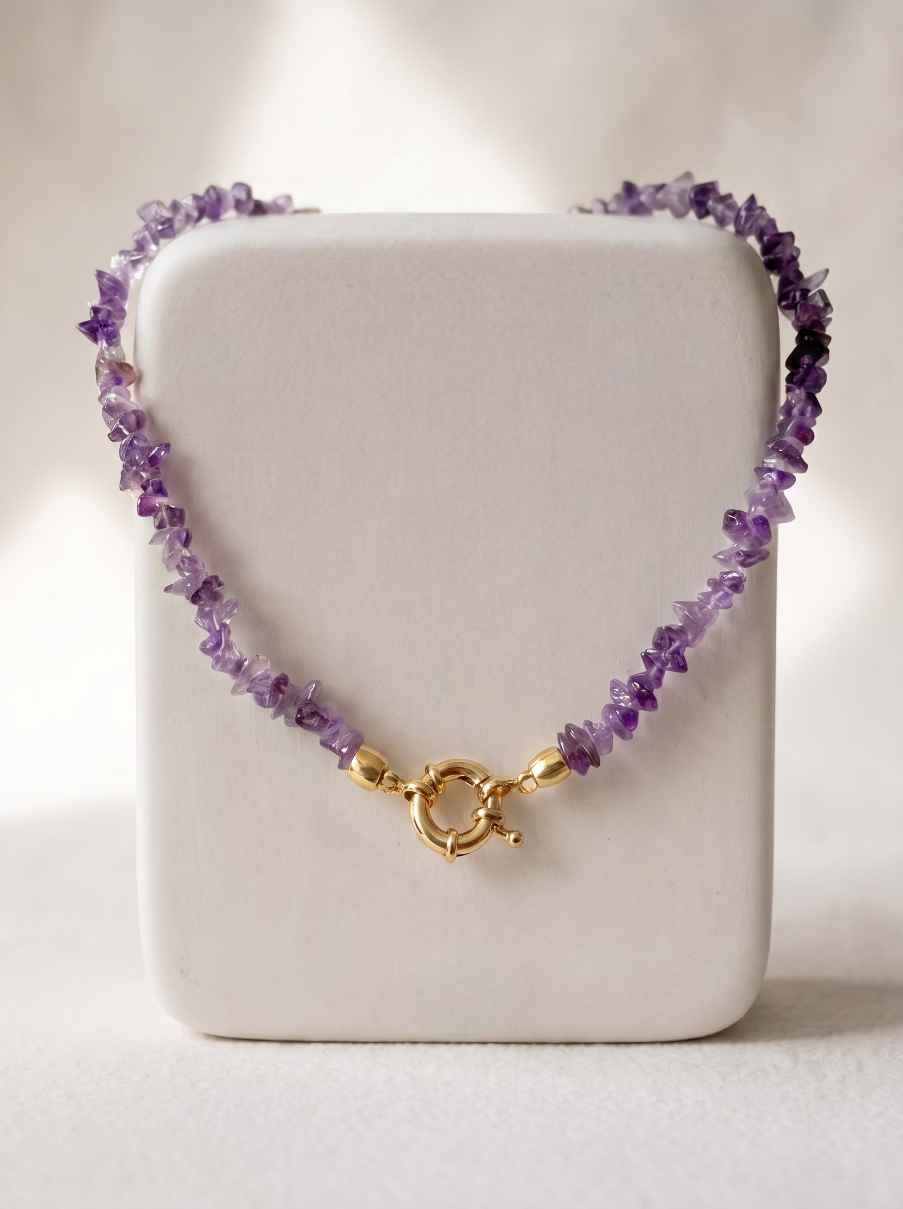 A beaded necklace made of small purple amethyst chips with a gold clasp, displayed on a white rectangular jewelry stand against a softly lit neutral background.