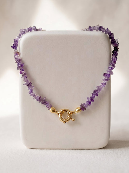 A beaded necklace made of small purple amethyst chips with a gold clasp, displayed on a white rectangular jewelry stand against a softly lit neutral background.