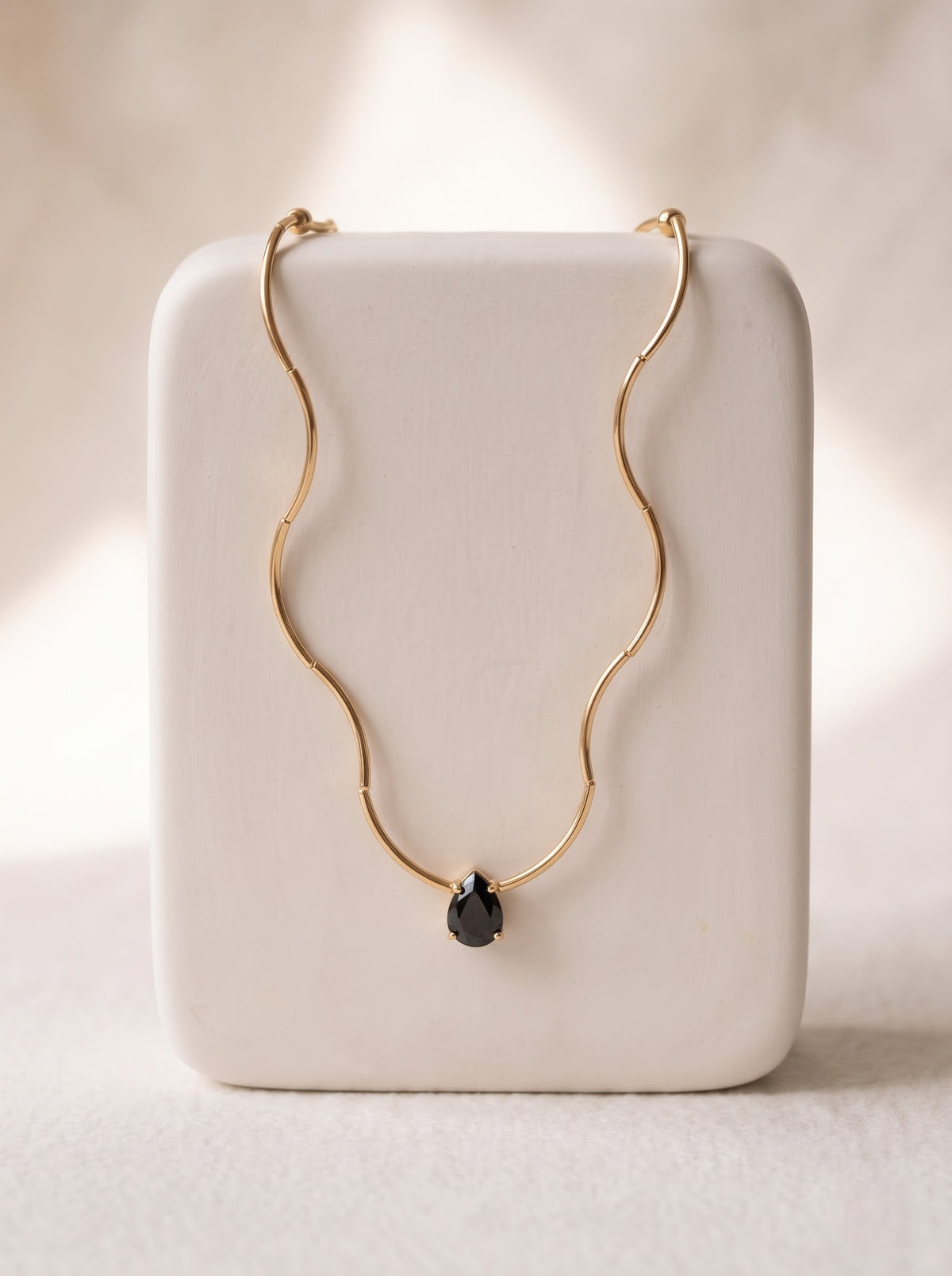 Woman in a white one-shoulder top wearing a StoneCraft Jewellery gold wave chain necklace with a black teardrop crystal pendant.