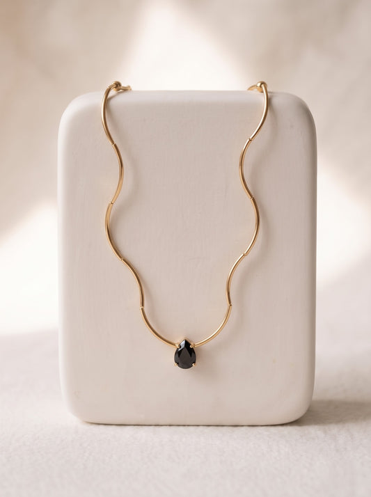 Woman in a white one-shoulder top wearing a StoneCraft Jewellery gold wave chain necklace with a black teardrop crystal pendant.