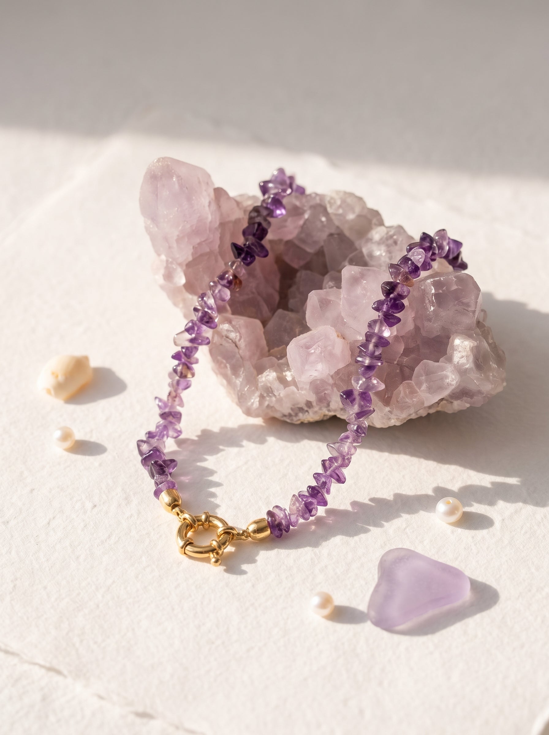 Necklace made of small purple gemstone chips and a gold clasp, displayed on a pale pink crystal cluster with a few small pearls and a lavender stone on a light textured surface.