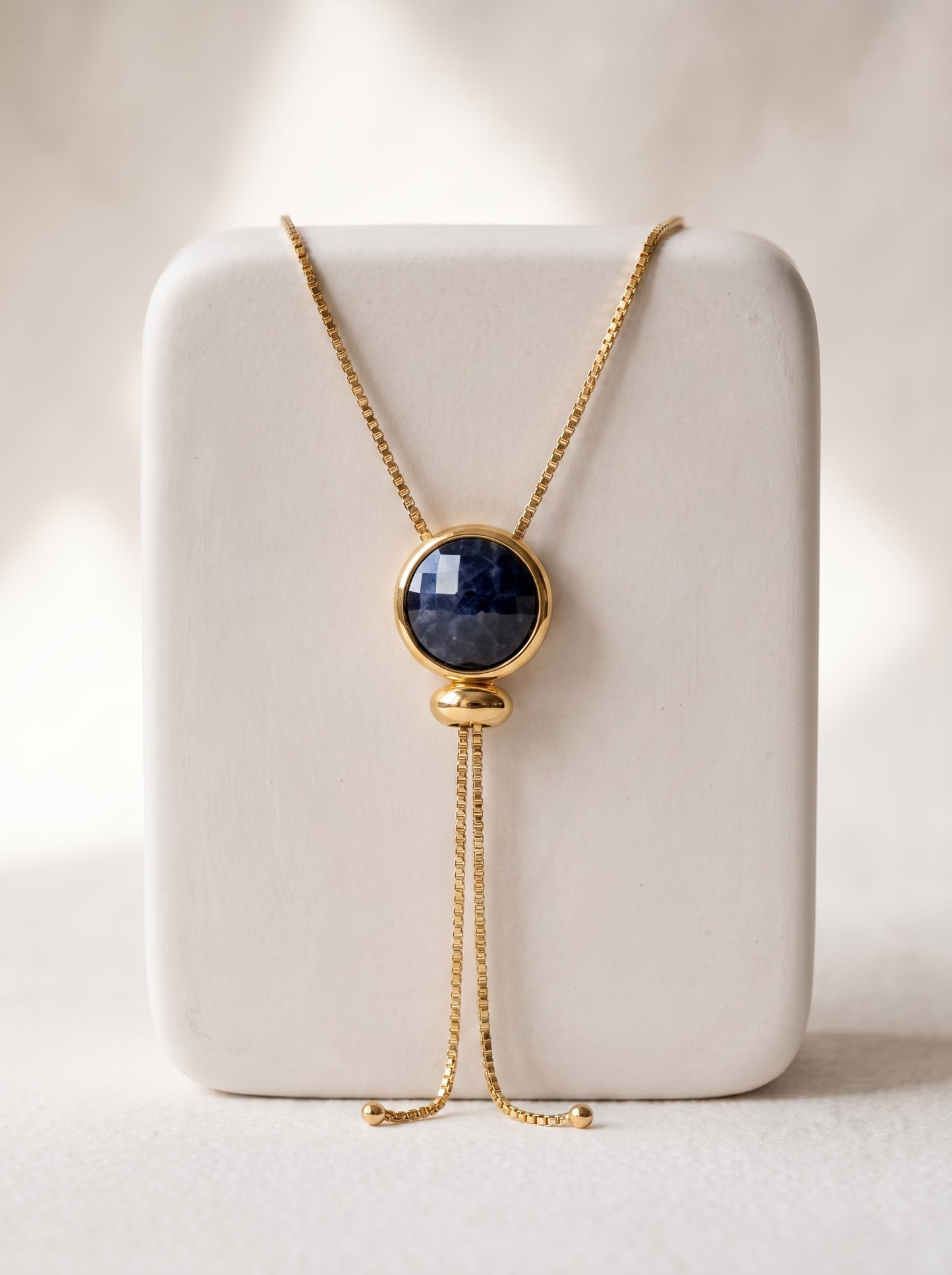 A gold-toned lariat necklace with a round dark blue  quartz cabochon pendant, displayed on a white rectangular jewelry stand against a soft beige background.