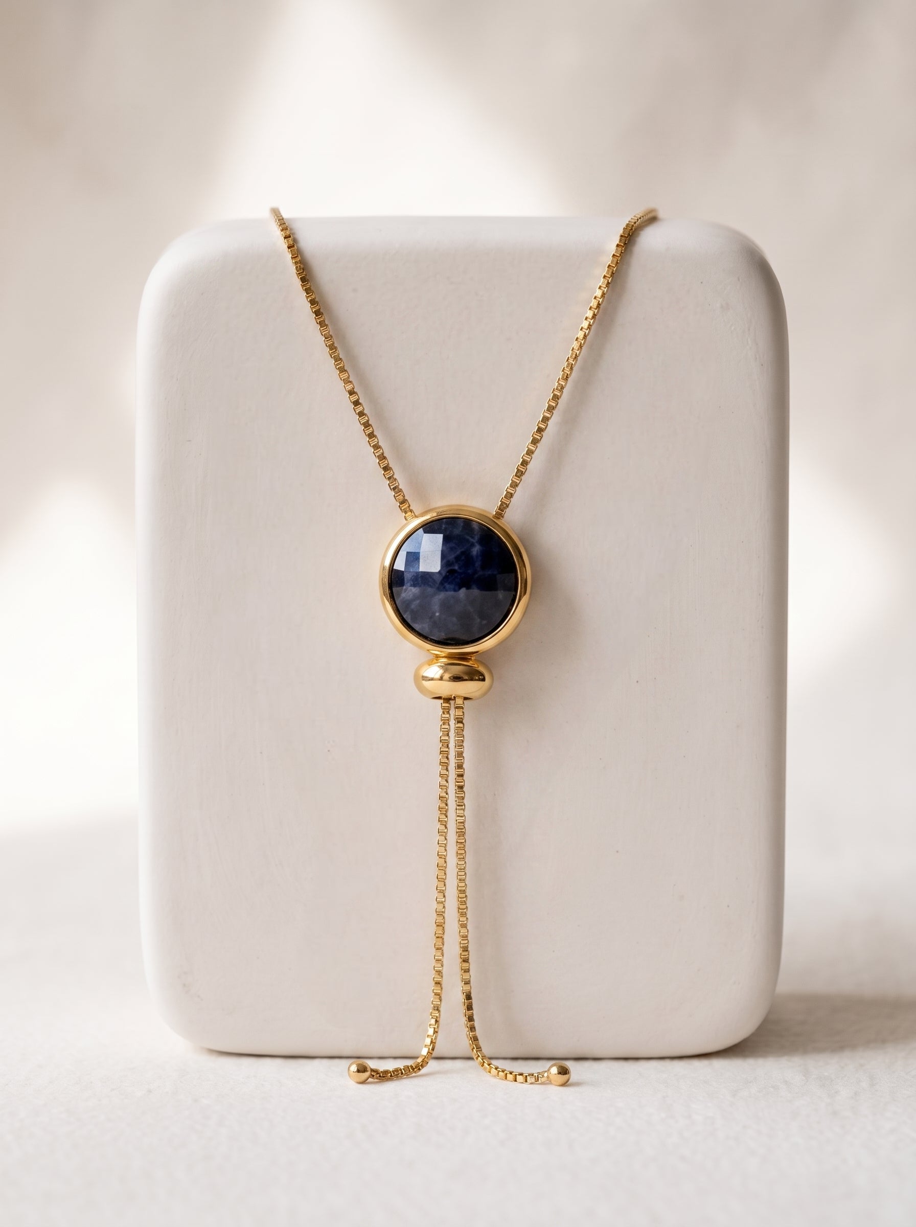 A gold-toned lariat necklace with a round dark blue  quartz cabochon pendant, displayed on a white rectangular jewelry stand against a soft beige background.