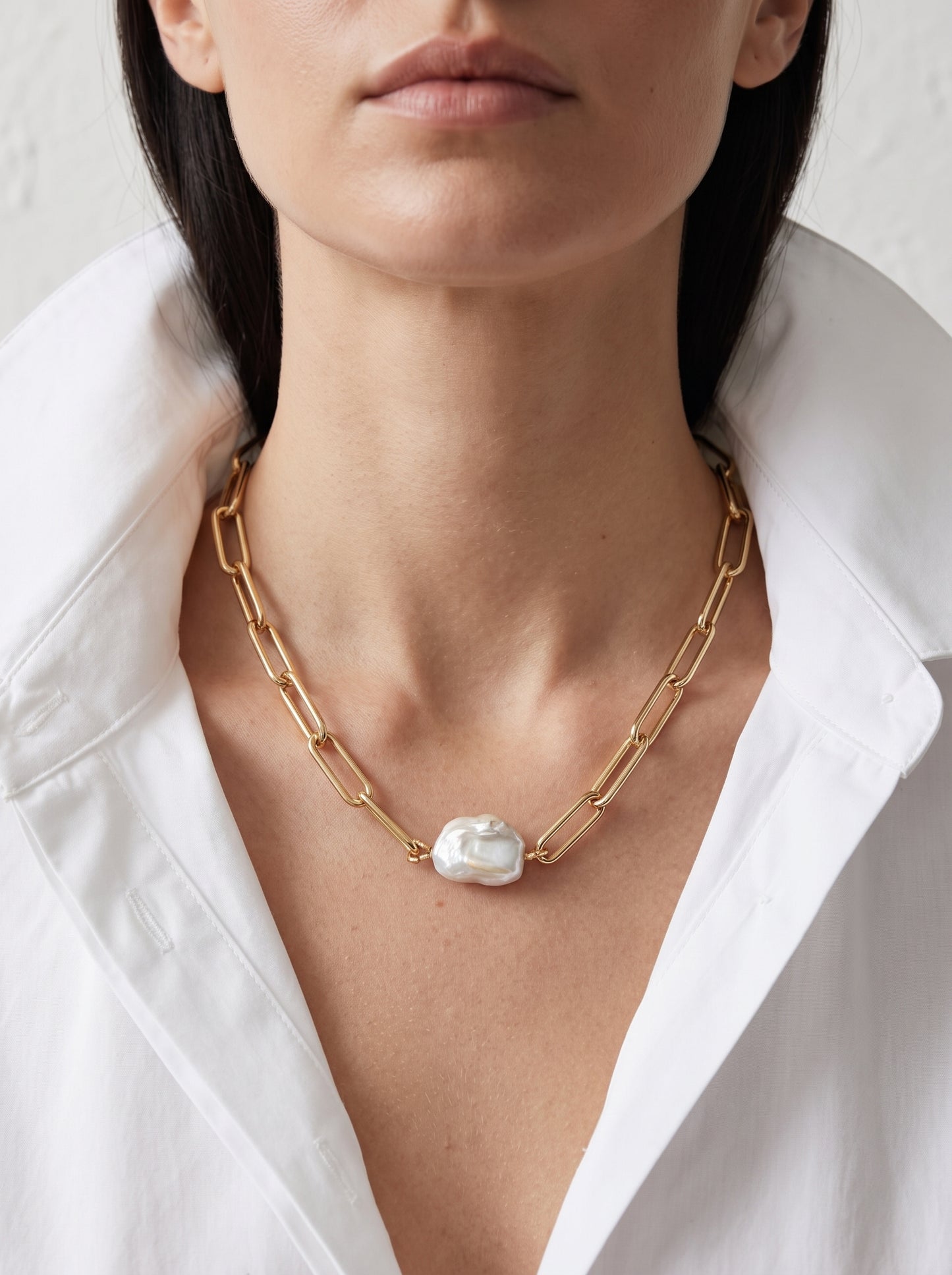A close-up of a model wearing a gold chain necklace with a large, irregular white pearl pendant, styled with a white collared shirt.