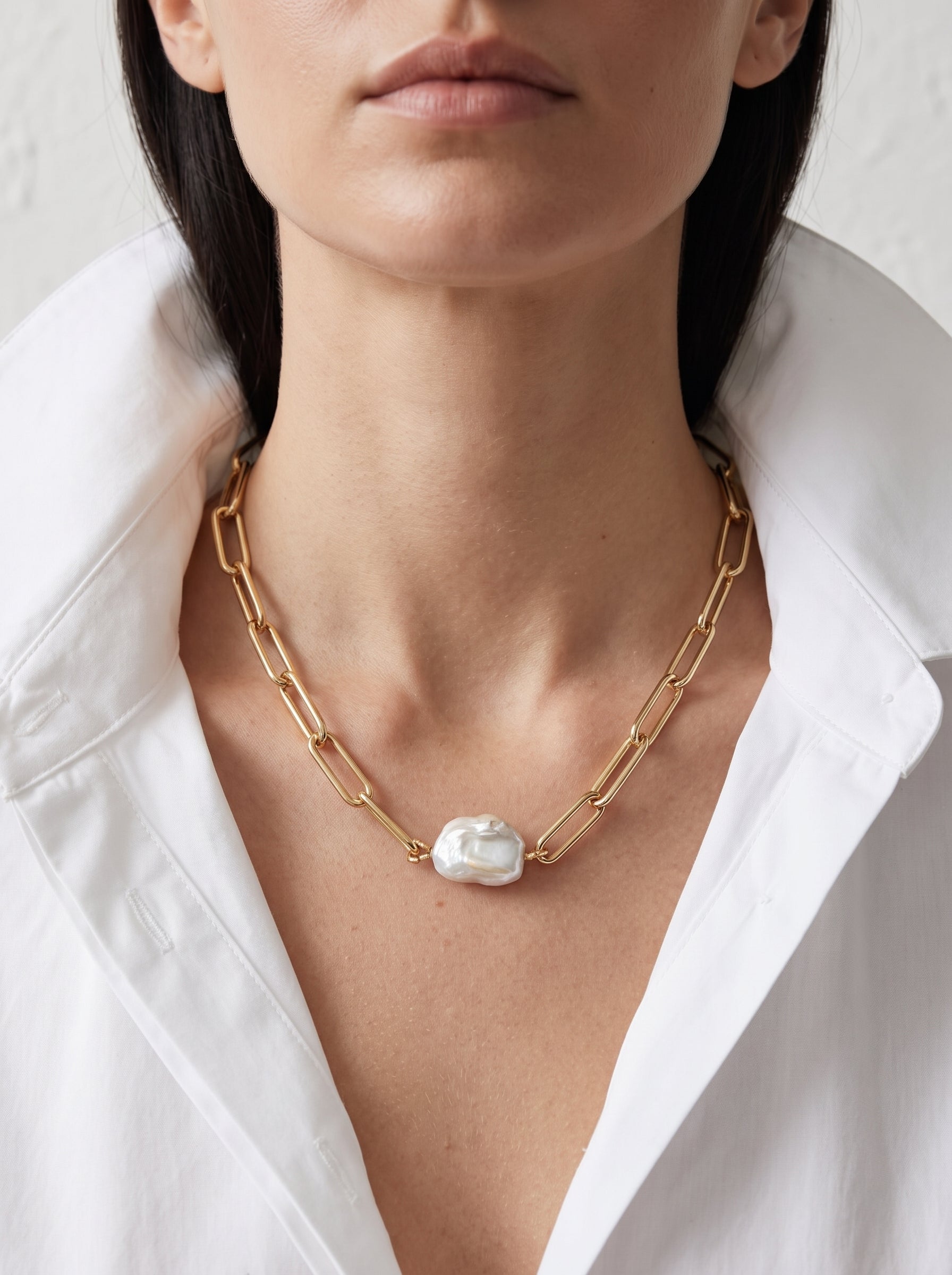A close-up of a model wearing a gold chain necklace with a large, irregular white pearl pendant, styled with a white collared shirt.
