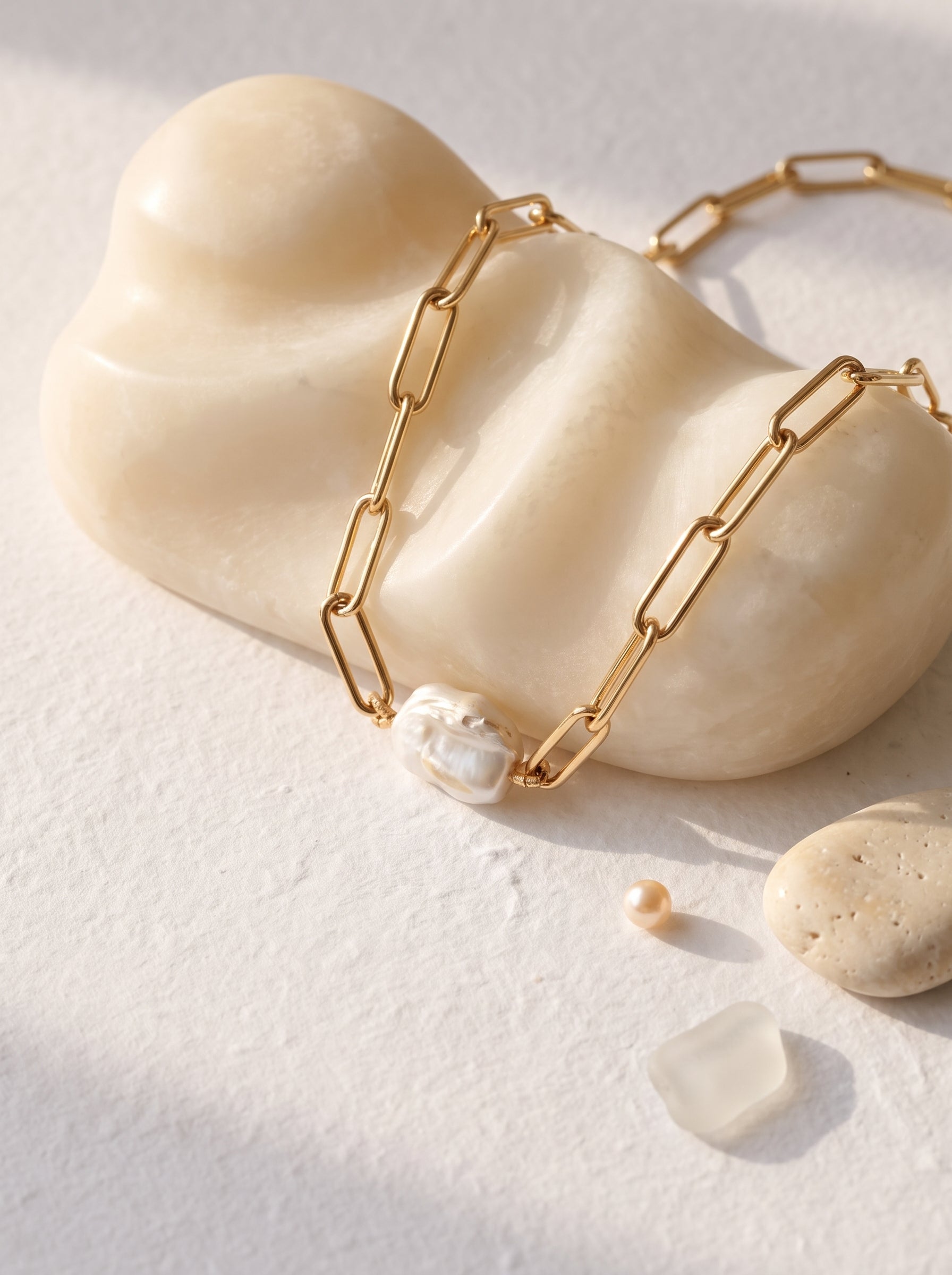A gold paperclip chain necklace with a small white pearl pendant, displayed on a cream-colored stone mannequin against a textured white background with natural shadows.