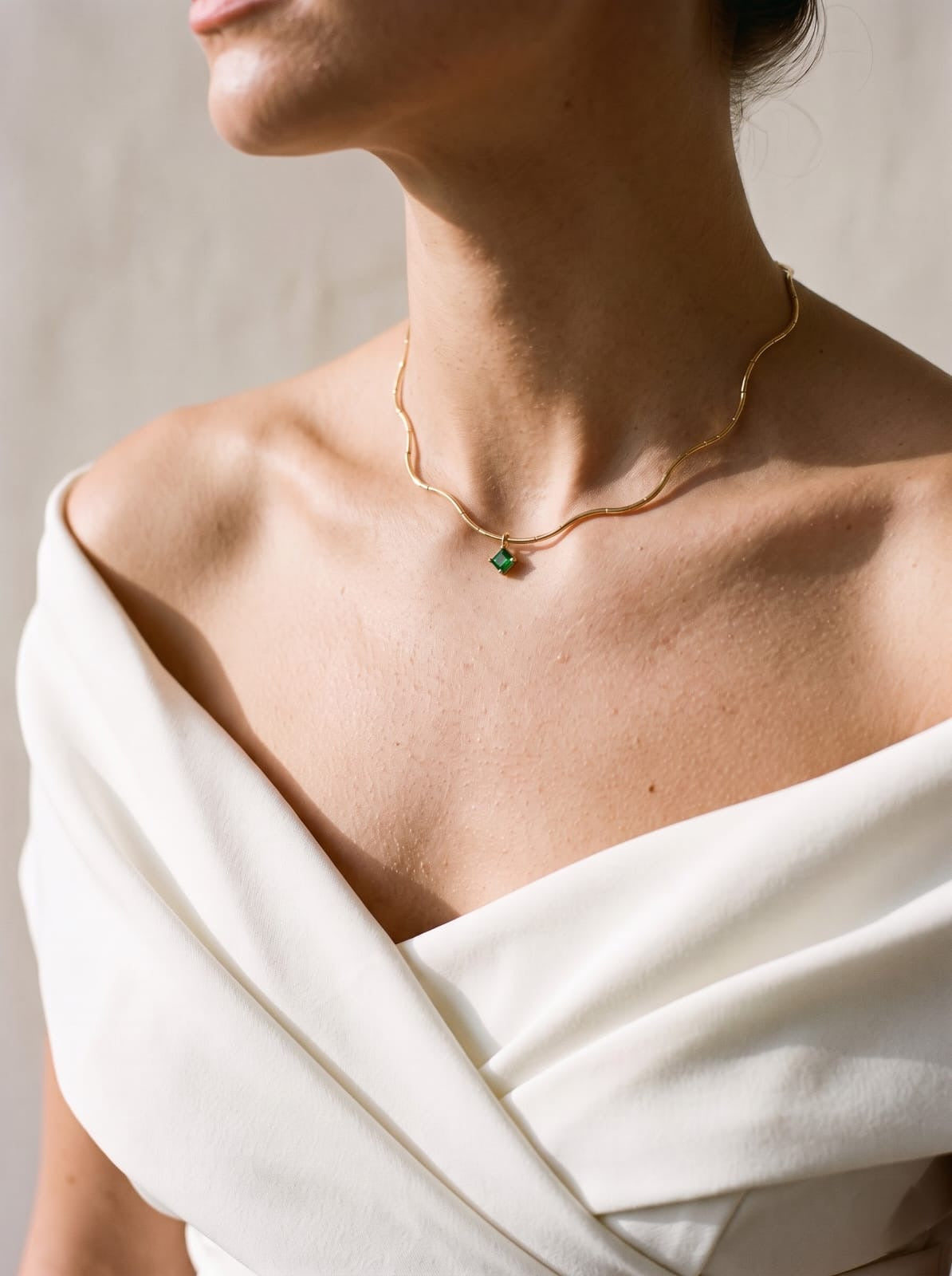A close-up lifestyle shot of a model wearing a delicate gold chain necklace with a small green gemstone pendant, styled with an off-the-shoulder white dress against a neutral background.