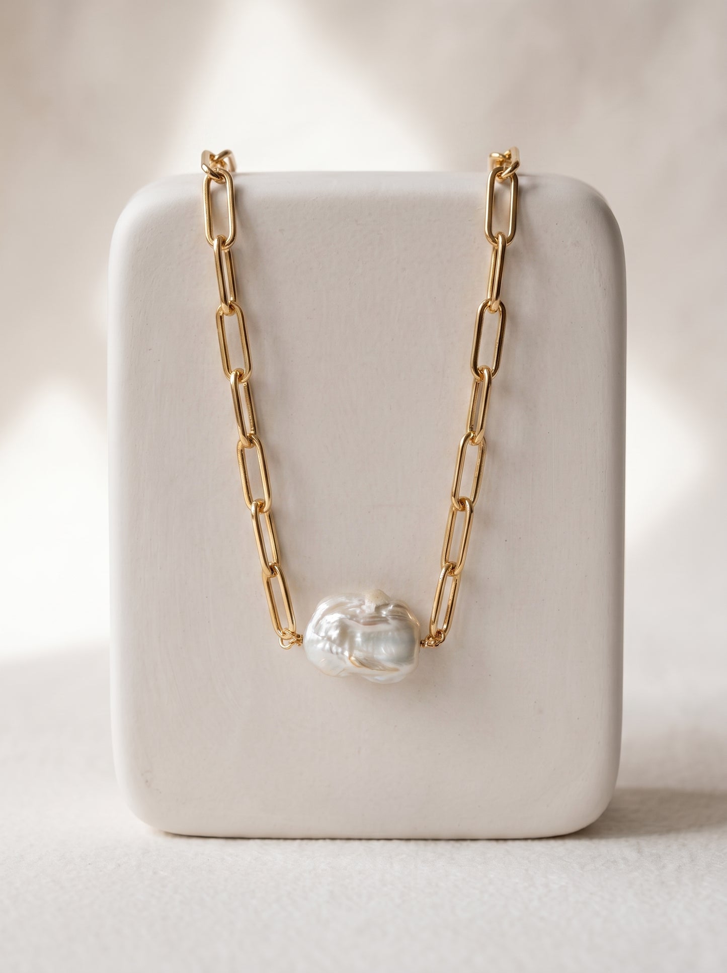 A gold paperclip chain necklace with a single irregular white pearl pendant, displayed on a white rectangular jewelry stand against a softly lit neutral background.