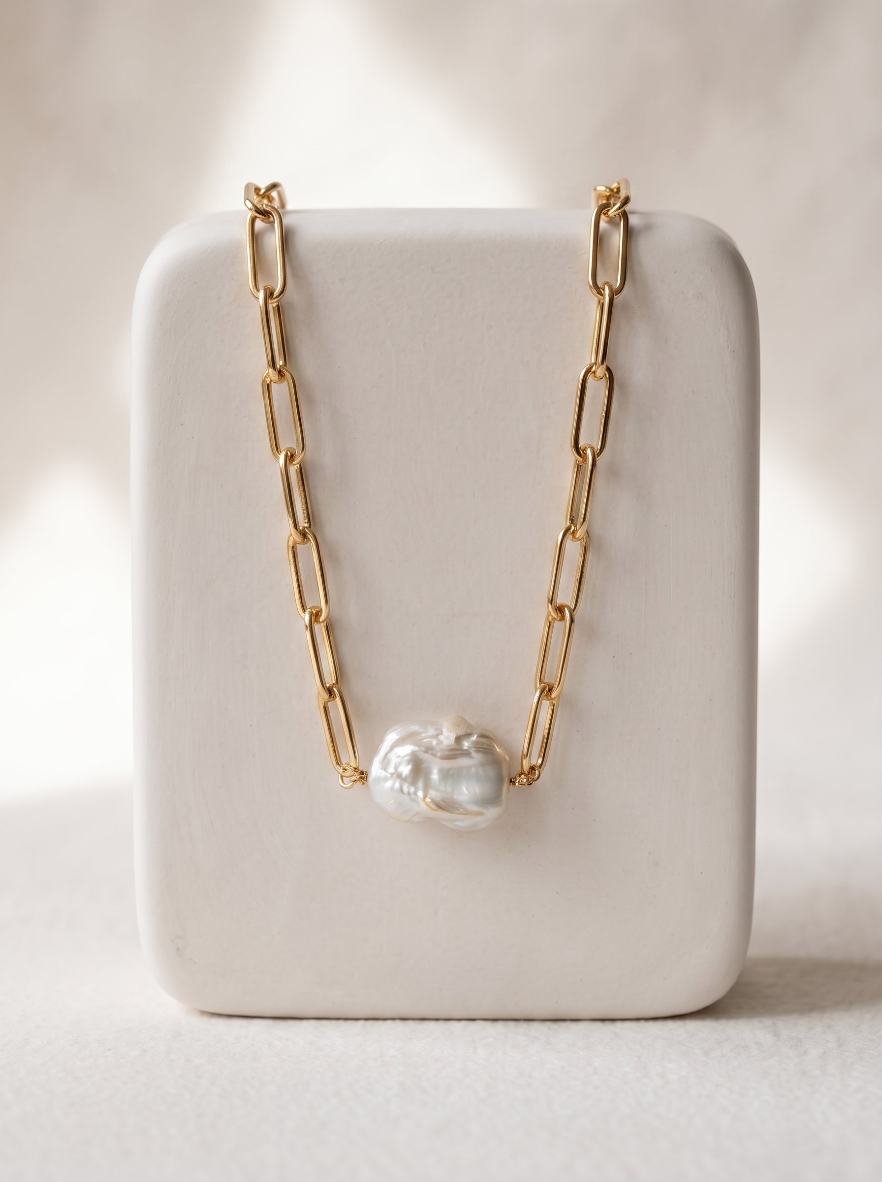A gold paperclip chain necklace with a single irregular white pearl pendant, displayed on a white rectangular jewelry stand against a softly lit neutral background.