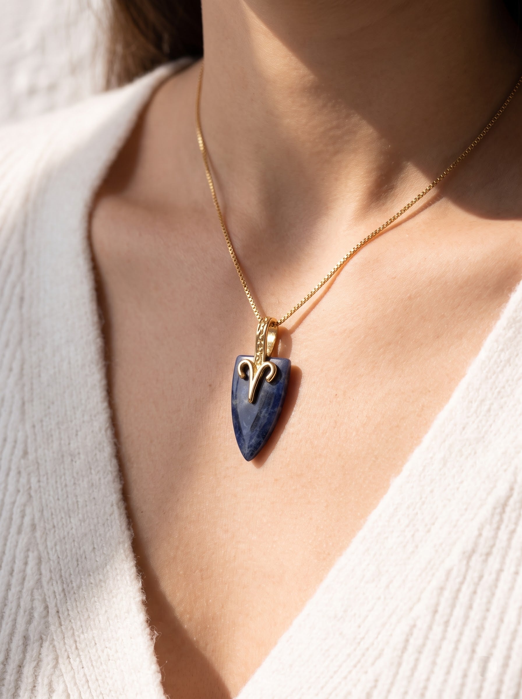 Woman wearing StoneCraft Jewellery sodalite Aries zodiac arrowhead pendant necklace on gold box chain, white knit top close-up.