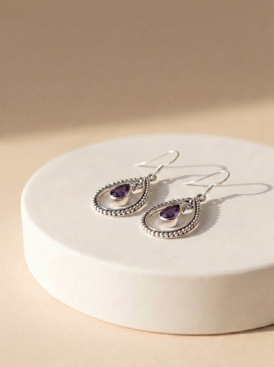 A pair of silver teardrop dangle earrings with textured beaded borders and purple amethyst centers, displayed on a round white pedestal against a warm beige background.