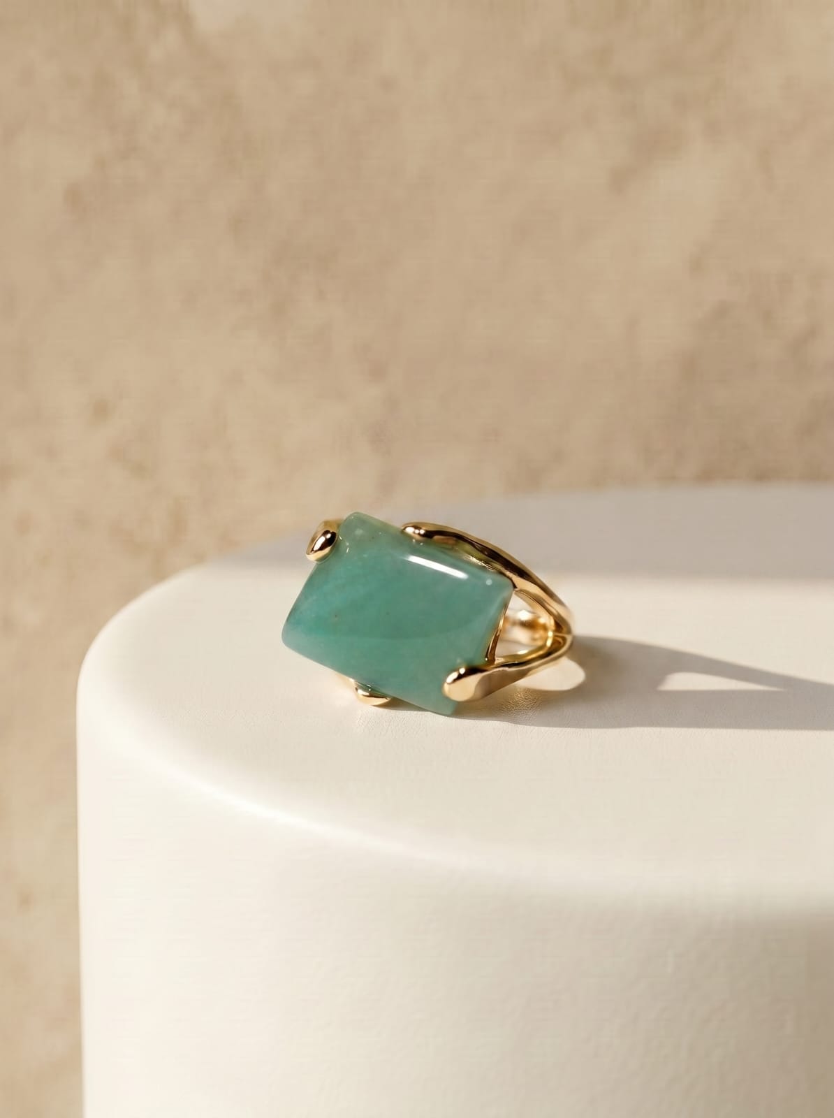 Natural green quartz statement ring with rectangular stone on gold-plated band, resting on a white curved surface against a warm beige background