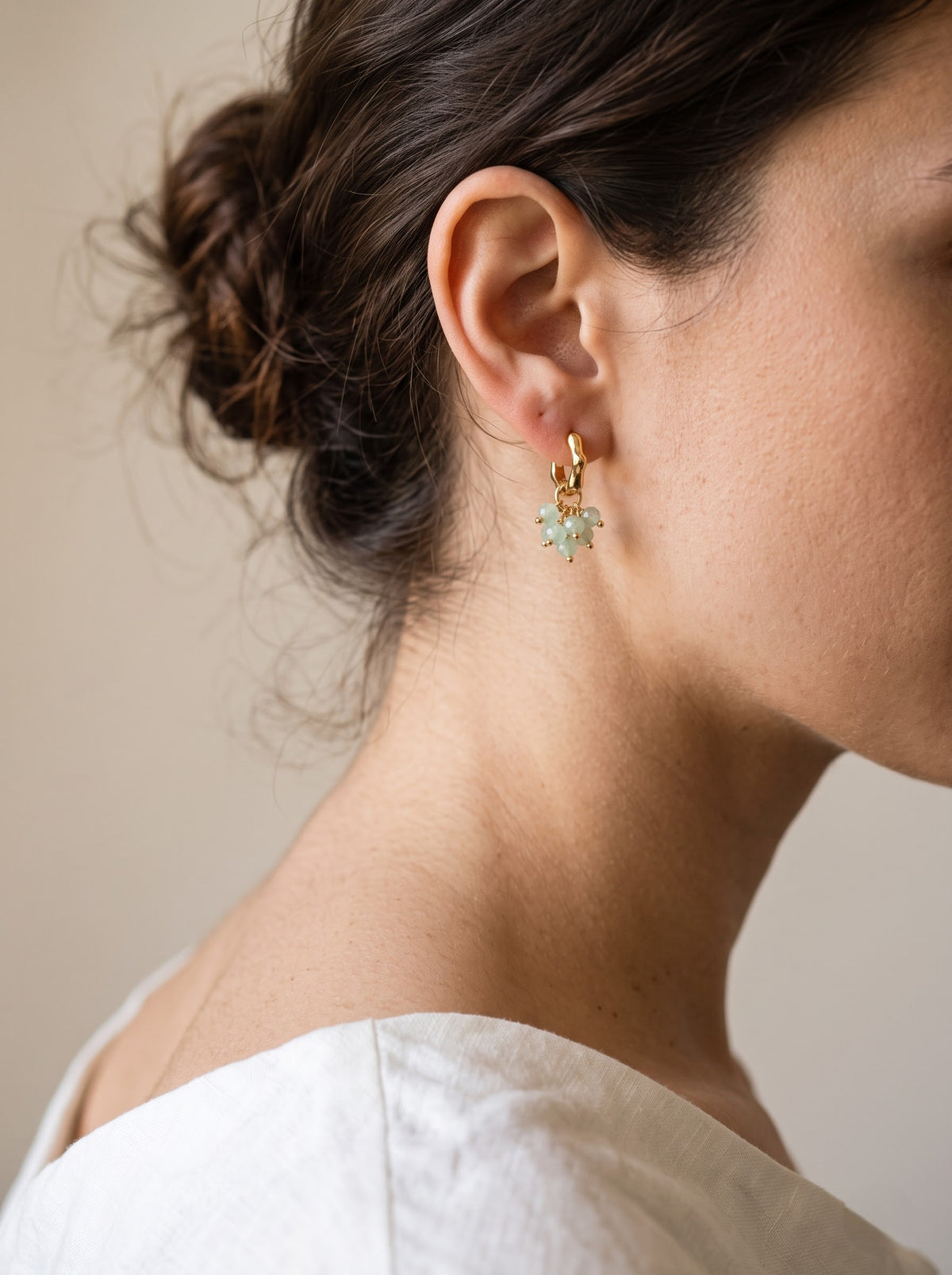 A close-up side profile of a woman wearing a delicate gold hoop earring with a small pale green aventurine dangle, against a soft beige background.