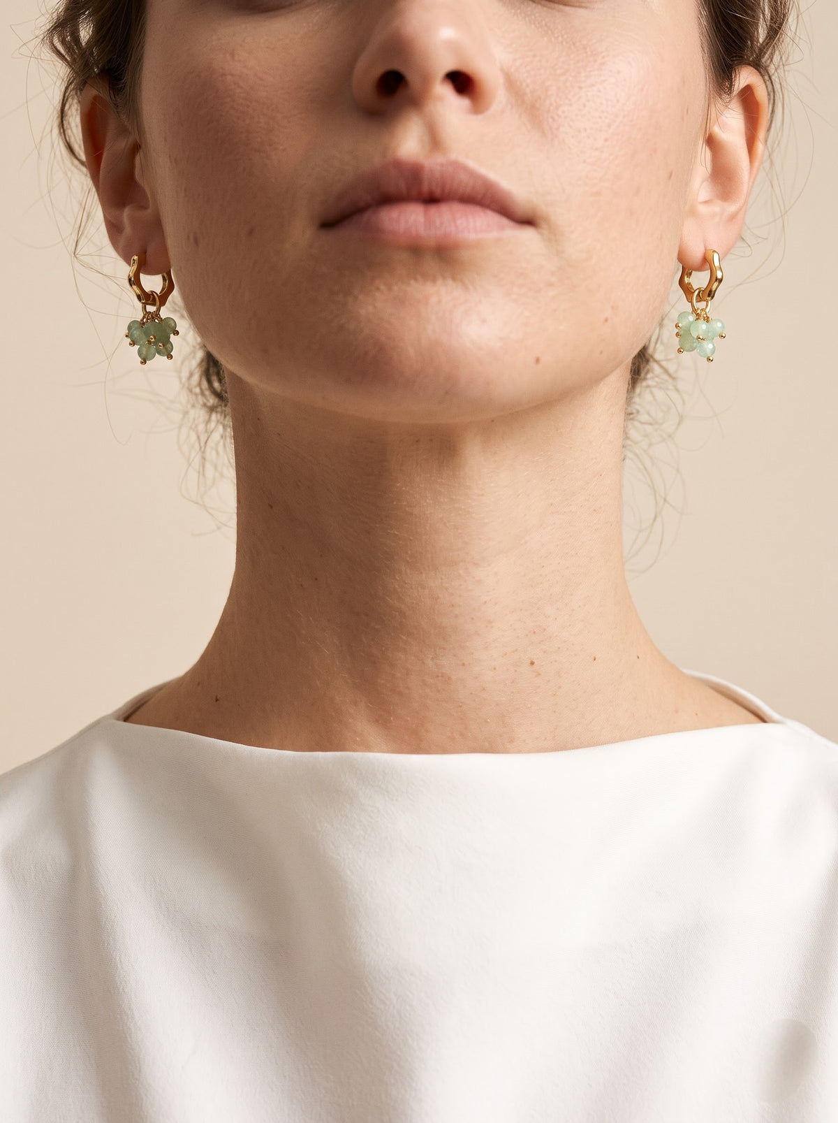 Close-up of a model wearing gold dangle earrings aventurine charms, paired with a white top against a warm beige background.