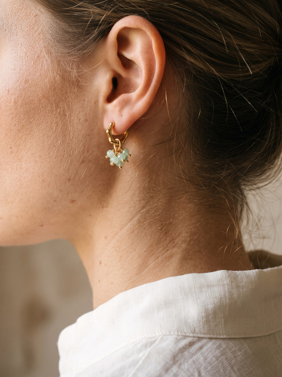 A close-up side profile of a woman wearing a delicate gold huggie hoop earring with a small dangling green aventurine charm.