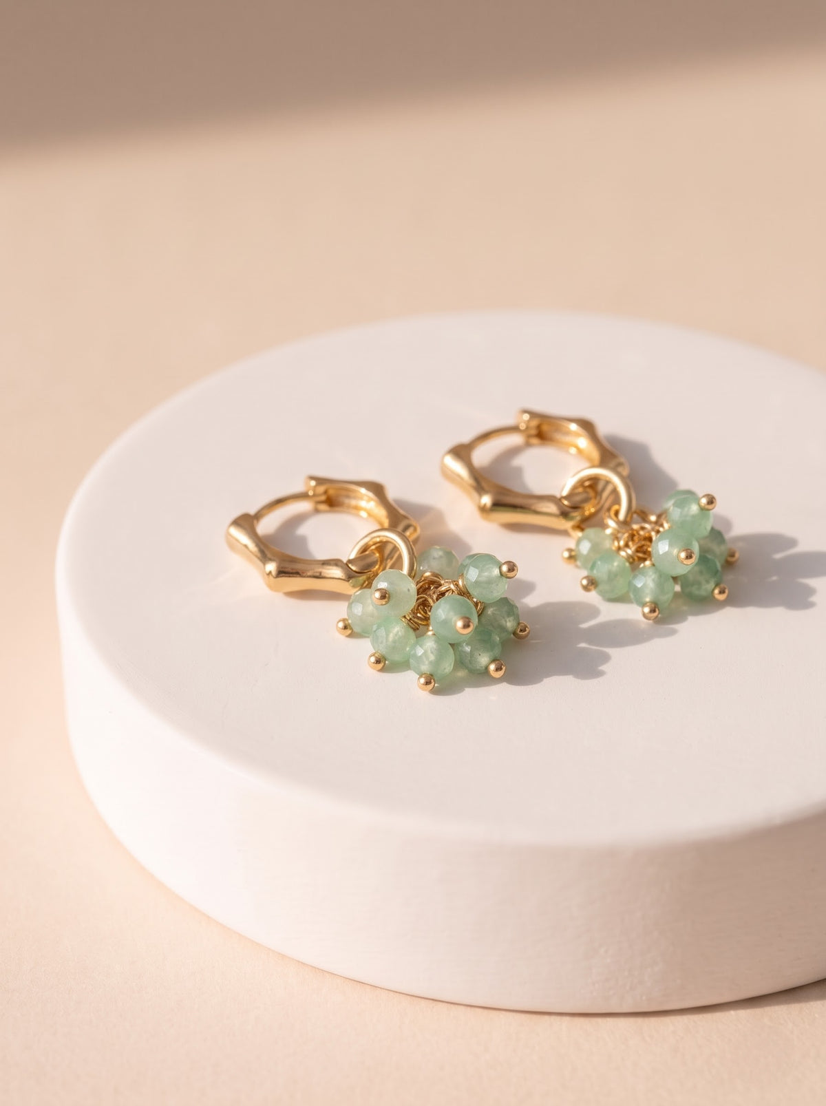 A pair of gold hoop earrings with dangling clusters of light green  aventurine faceted beads, displayed on a round white pedestal against a warm beige background.