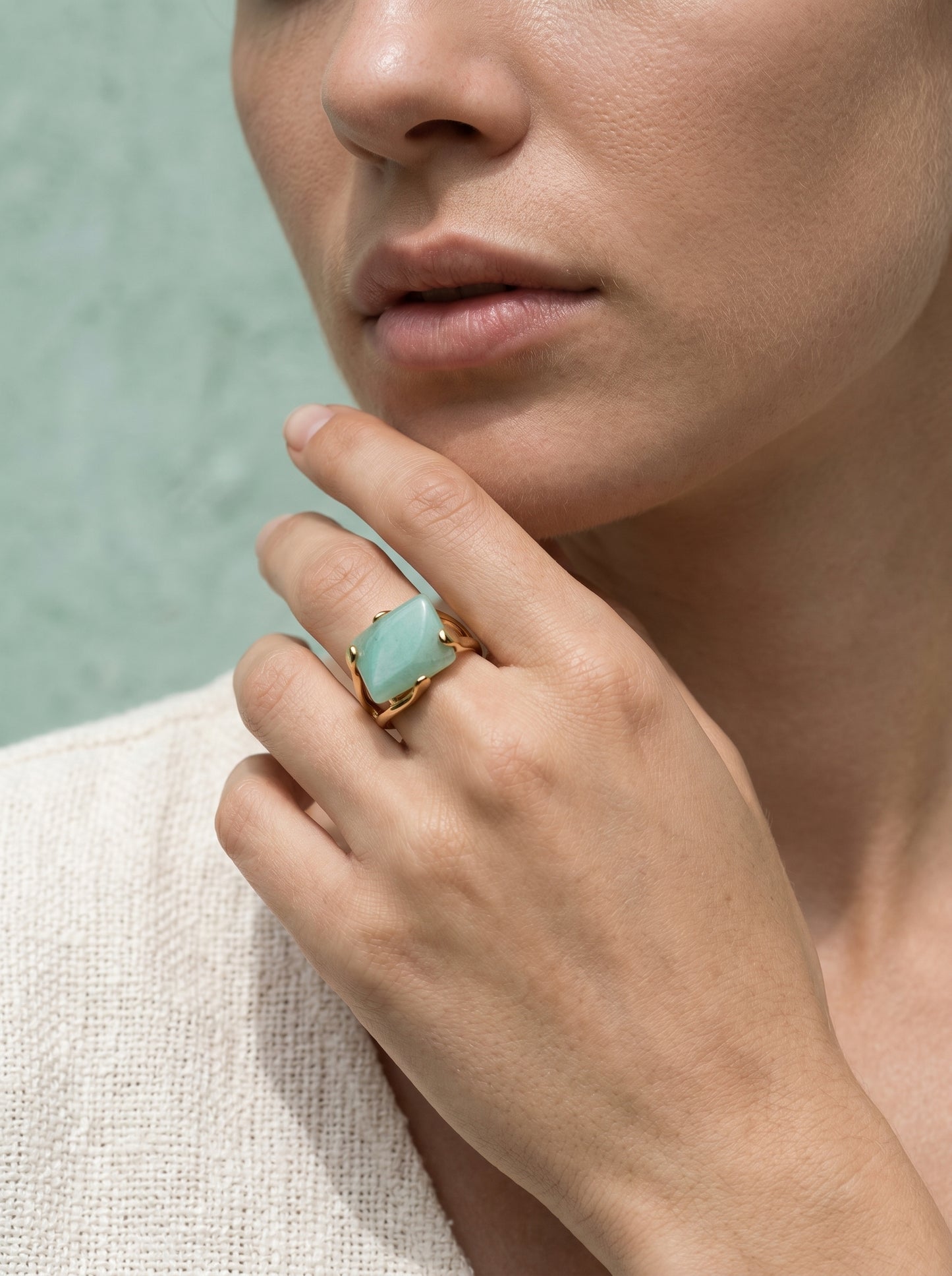Model wearing natural green quartz statement ring on gold-plated band, hand raised to chin, against a soft green background

