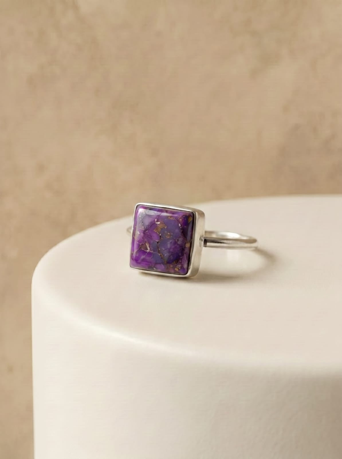 A silver ring with a square purple stone containing gold flecks, resting on a white cylindrical pedestal against a textured beige background.
