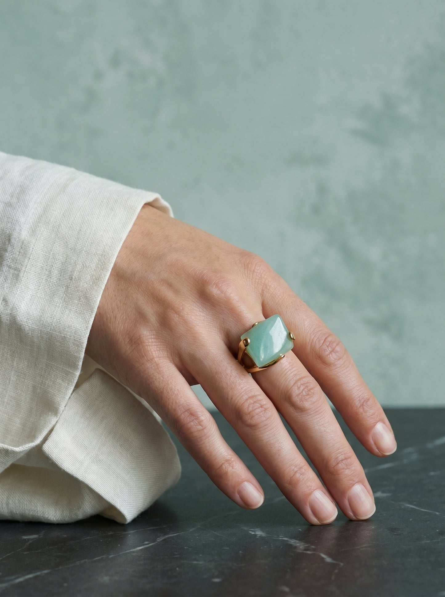 Model wearing natural green quartz statement ring on gold-plated band, hand raised to chin, against a soft green background

