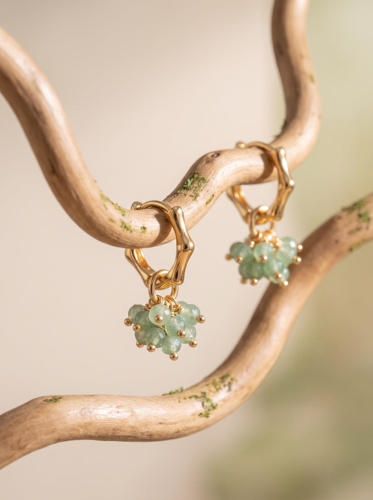 Gold-tone dangle earrings with clustered light green aventurine gemstones, hanging from a rustic curved wooden branch against a soft beige background.