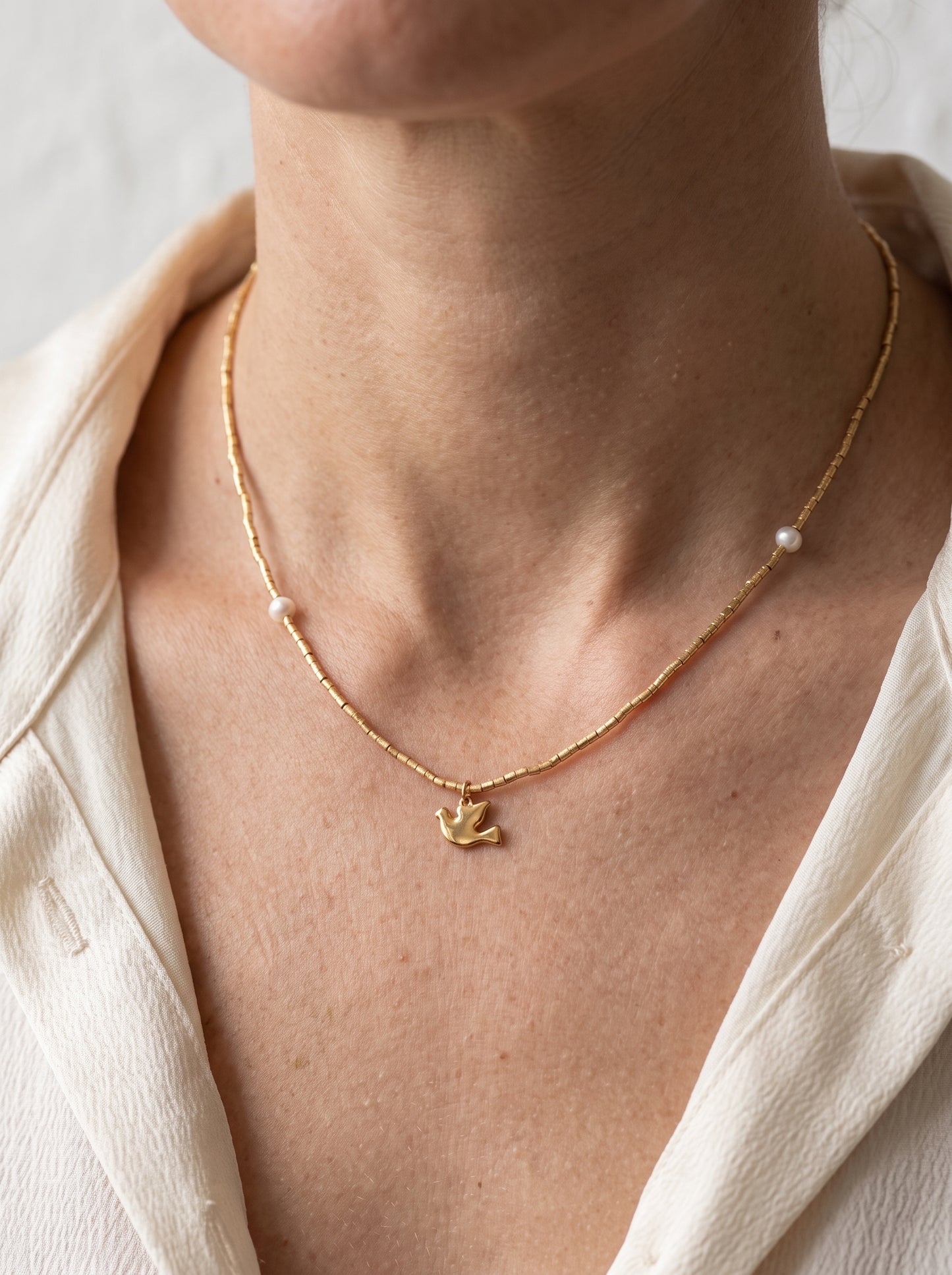 Gold plated beaded freshwater pearl necklace with bird charm worn on a woman's neckline against a cream blazer