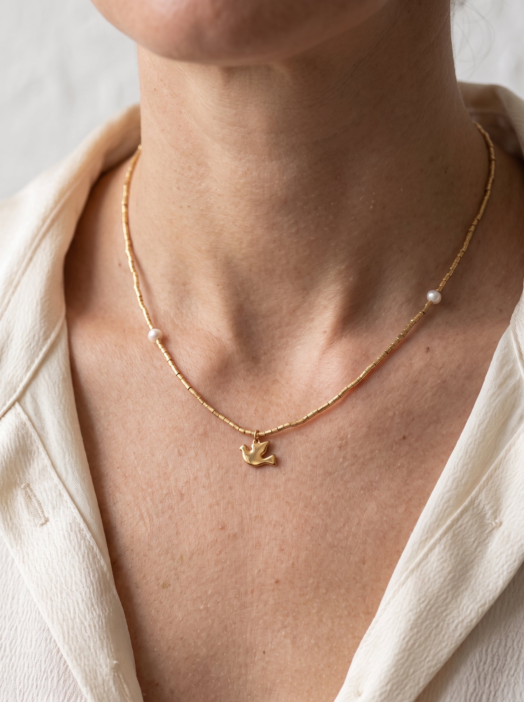 Gold plated beaded freshwater pearl necklace with bird charm worn on a woman's neckline against a cream blazer