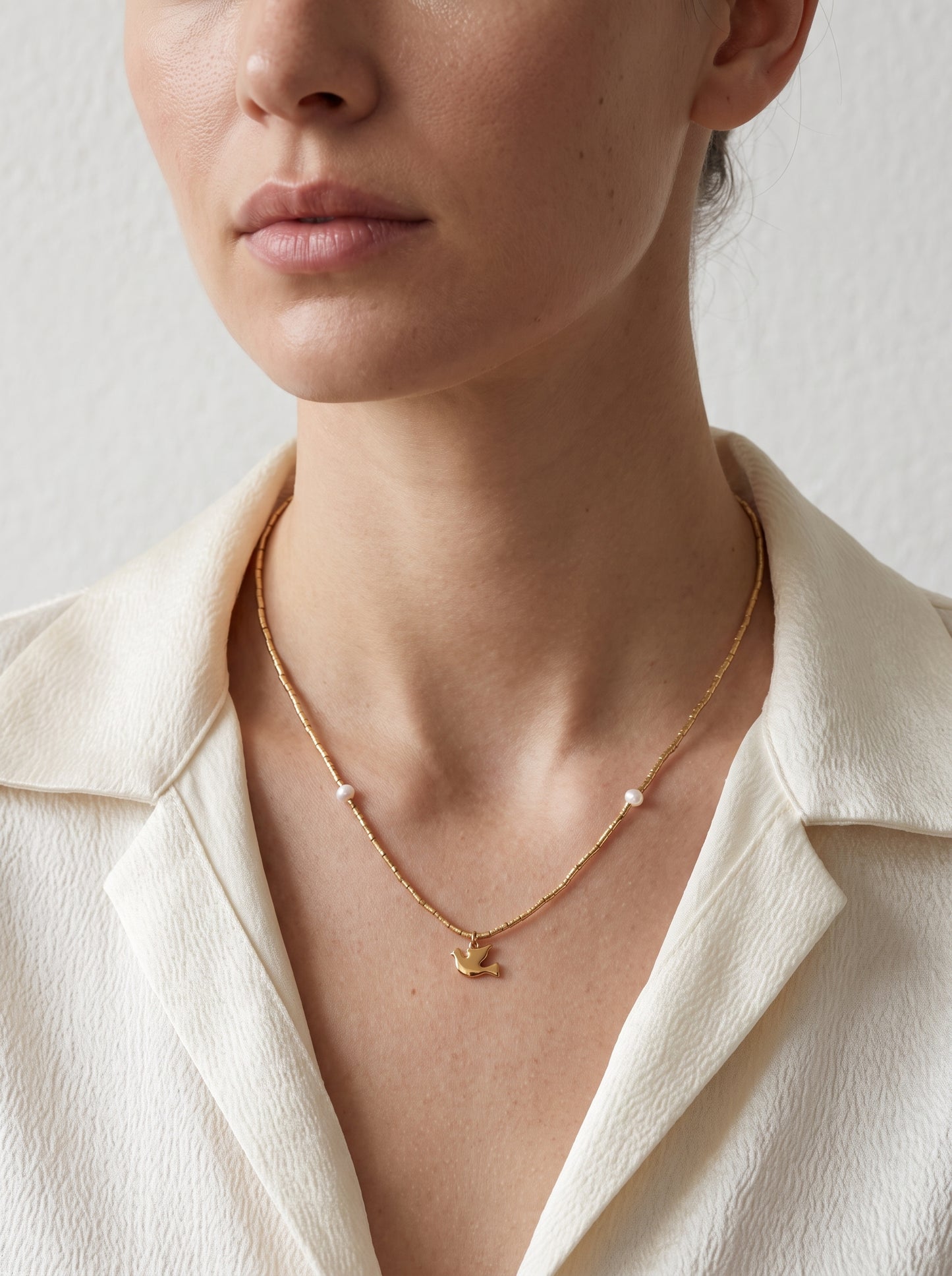 A close-up of a model wearing a delicate gold chain necklace with a small gold bird pendant and small white bead accents, styled with a cream-colored collared shirt.