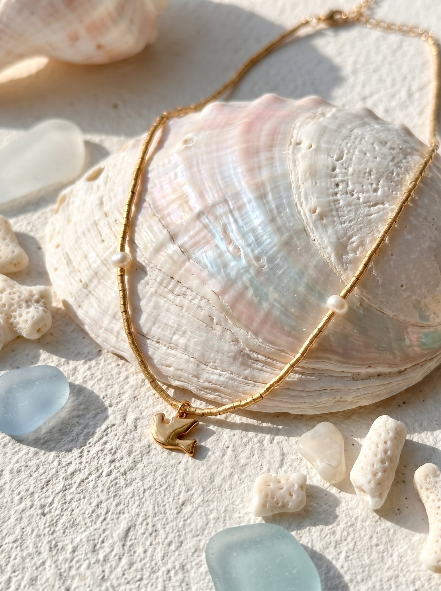 Gold-plated beaded pearl necklace with bird charm pendant displayed on an iridescent shell with sea glass and coral on sand