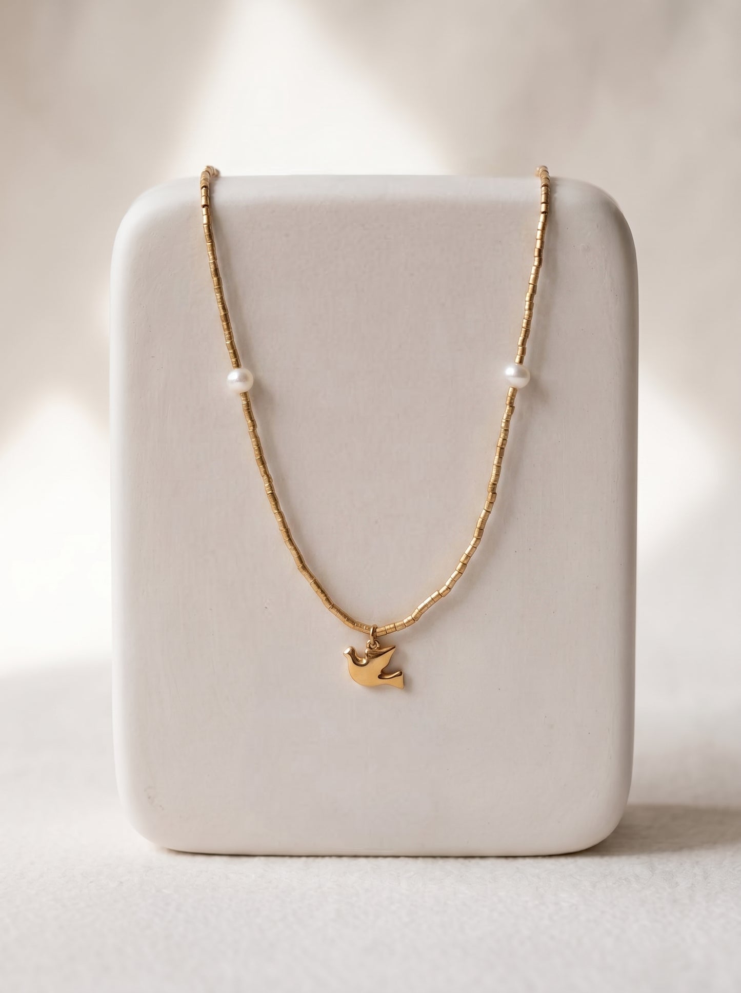 Gold-plated beaded necklace with two freshwater pearls and a gold flying bird charm pendant displayed on a white jewellery stand