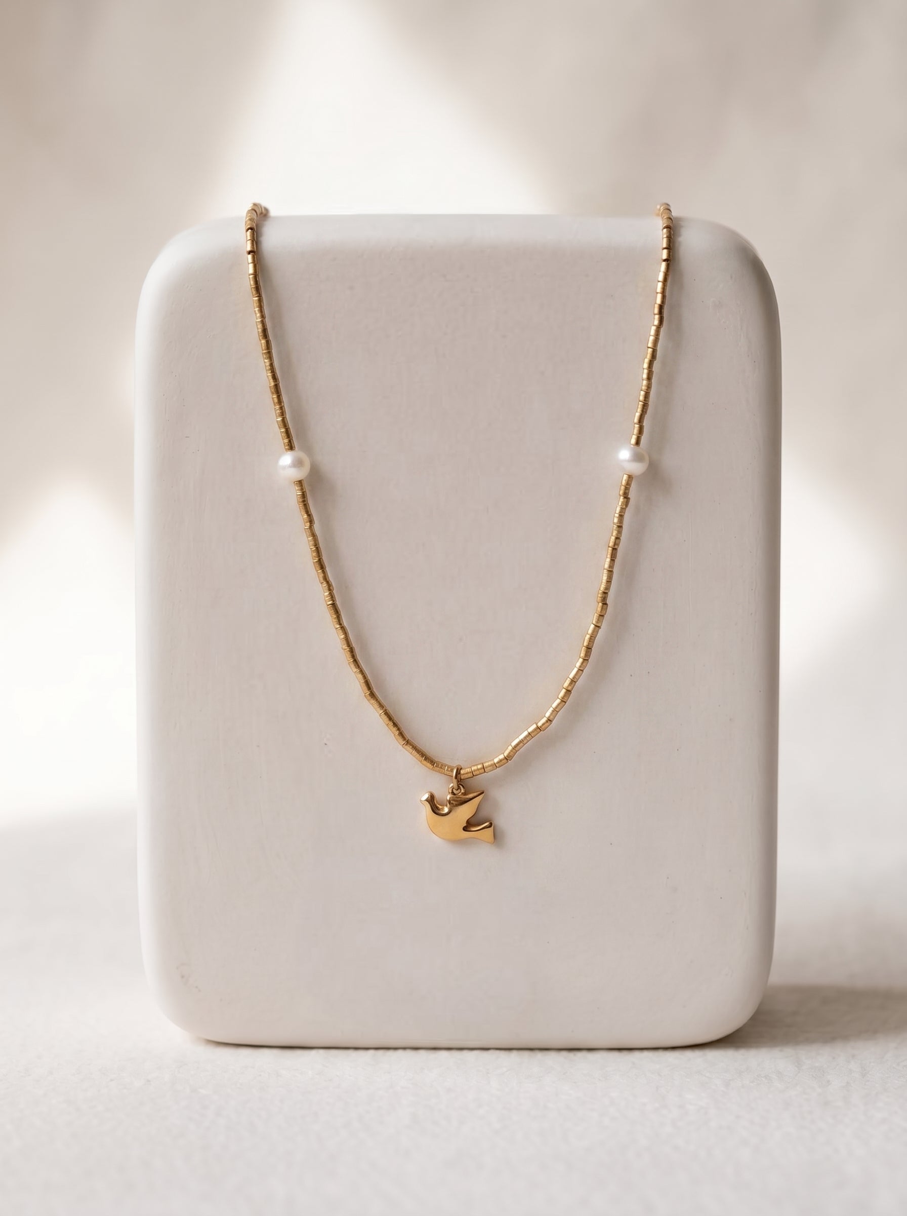 Gold-plated beaded necklace with two freshwater pearls and a gold flying bird charm pendant displayed on a white jewellery stand