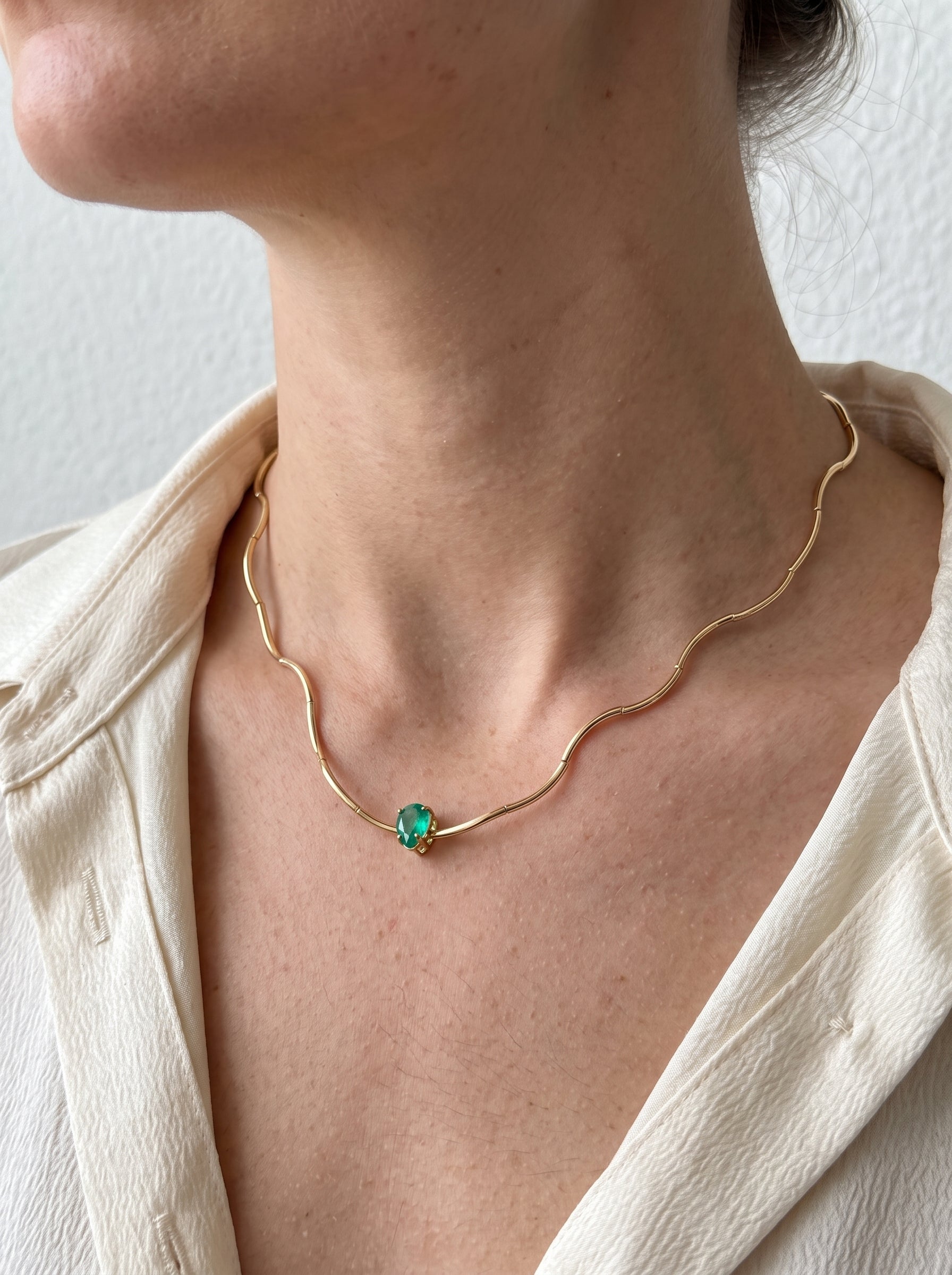 A close-up of a model wearing a delicate gold wavy chain necklace with a small green gemstone pendant, paired with a cream-colored linen shirt.
