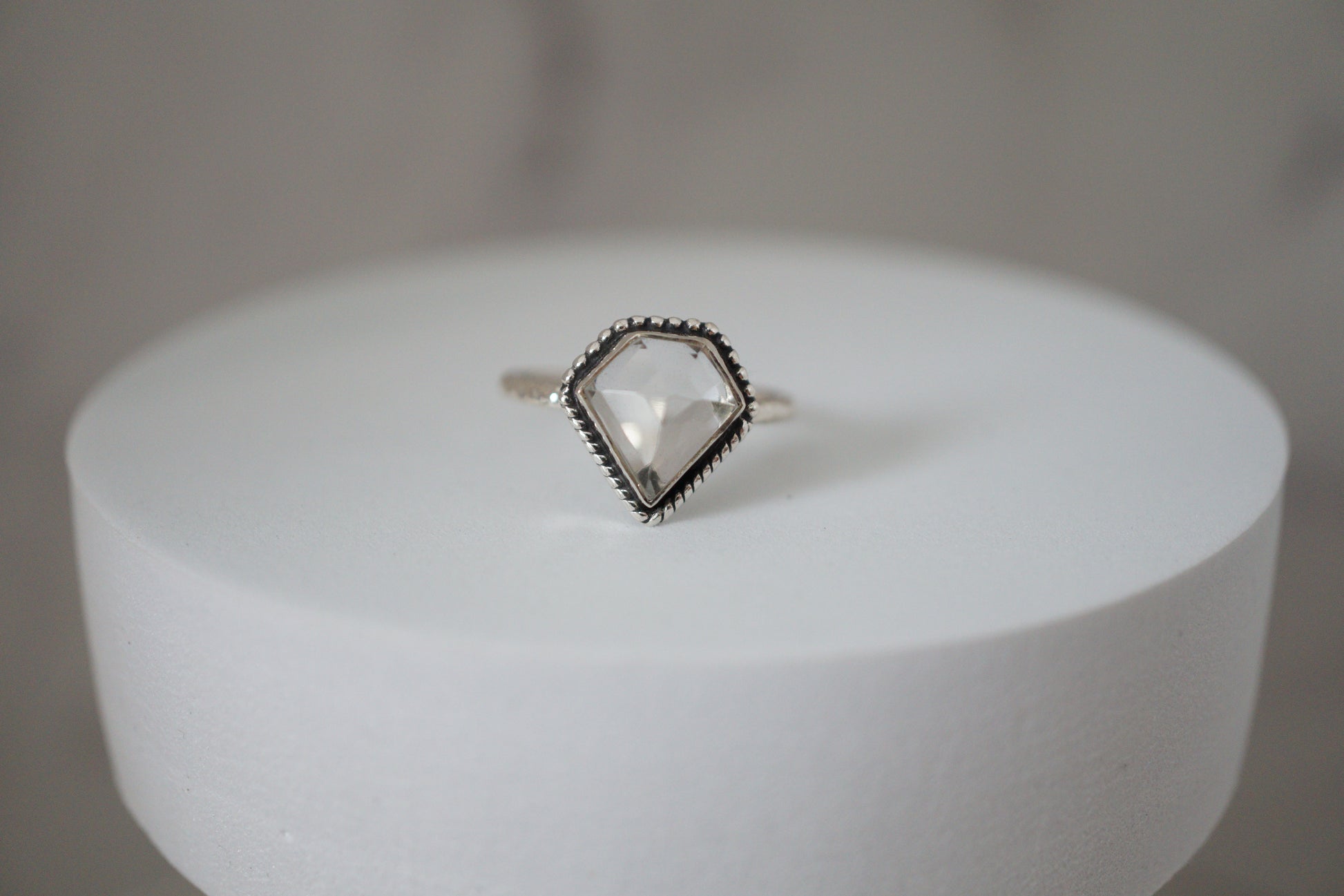 Ring with a clear  quartz gemstone on a white cylindrical stand against a gray background