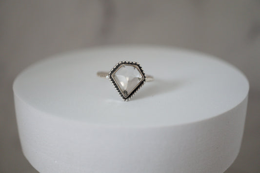Ring with a clear  quartz gemstone on a white cylindrical stand against a gray background
