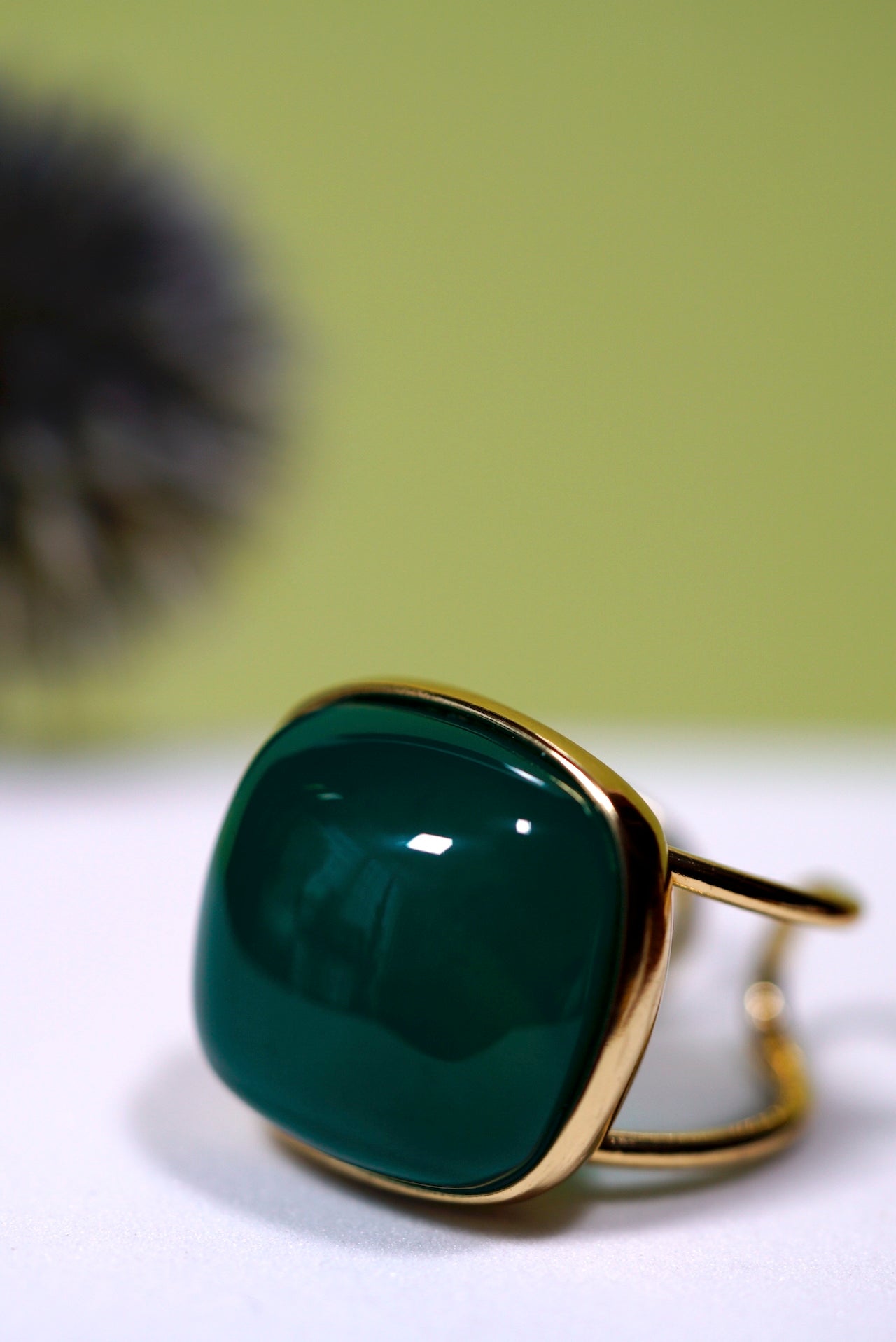 Green  cushion agate ring with gold  cuff band on a white surface with a blurred green background