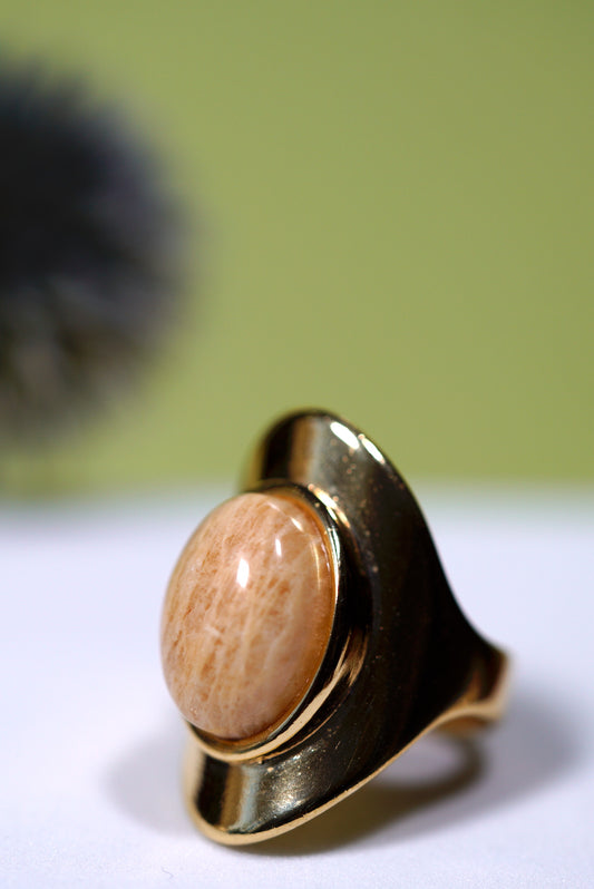 Gold ring with a beige amazonite  pear shape gemstone on a white surface with a blurred green background
