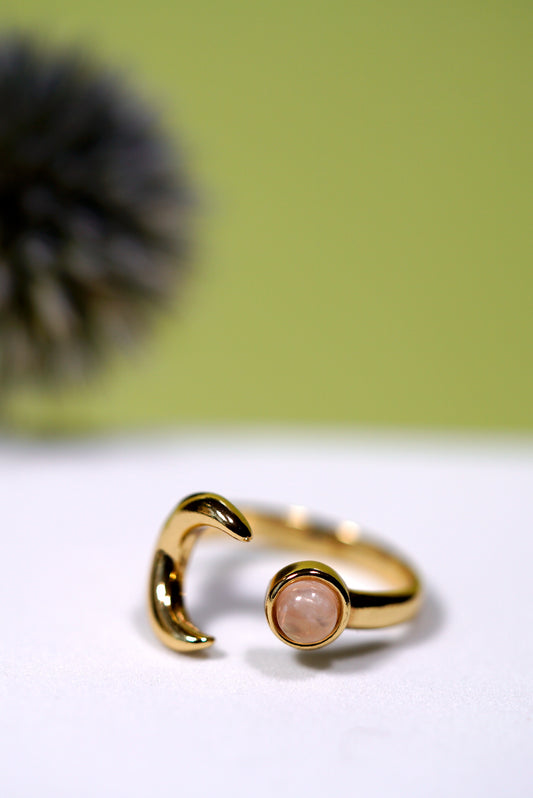 Gold ring with a moon and rose quartz gemstone on a white surface with a green background