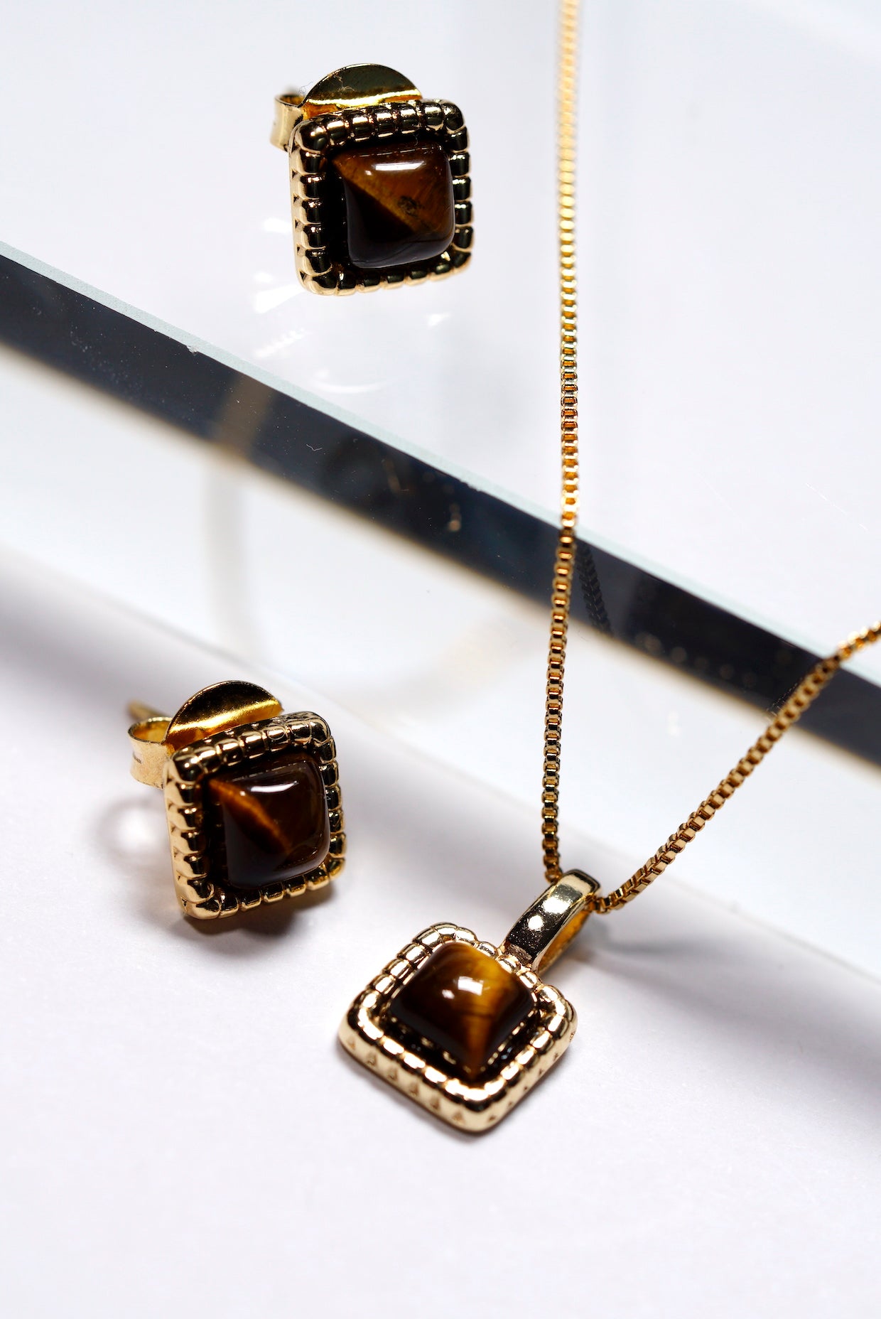 Jewellery set with gold chain, tigers eye hammered square dainty pendant, and tigers eye hammered square stud earrings on a reflective surface.