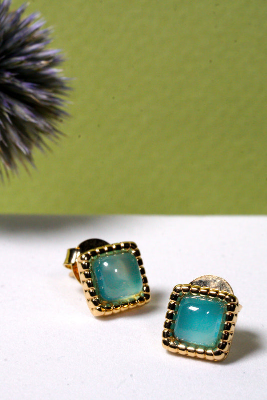Pair of gold-framed  hammered aqua agate gemstone square stud earrings on a white surface with a green background.