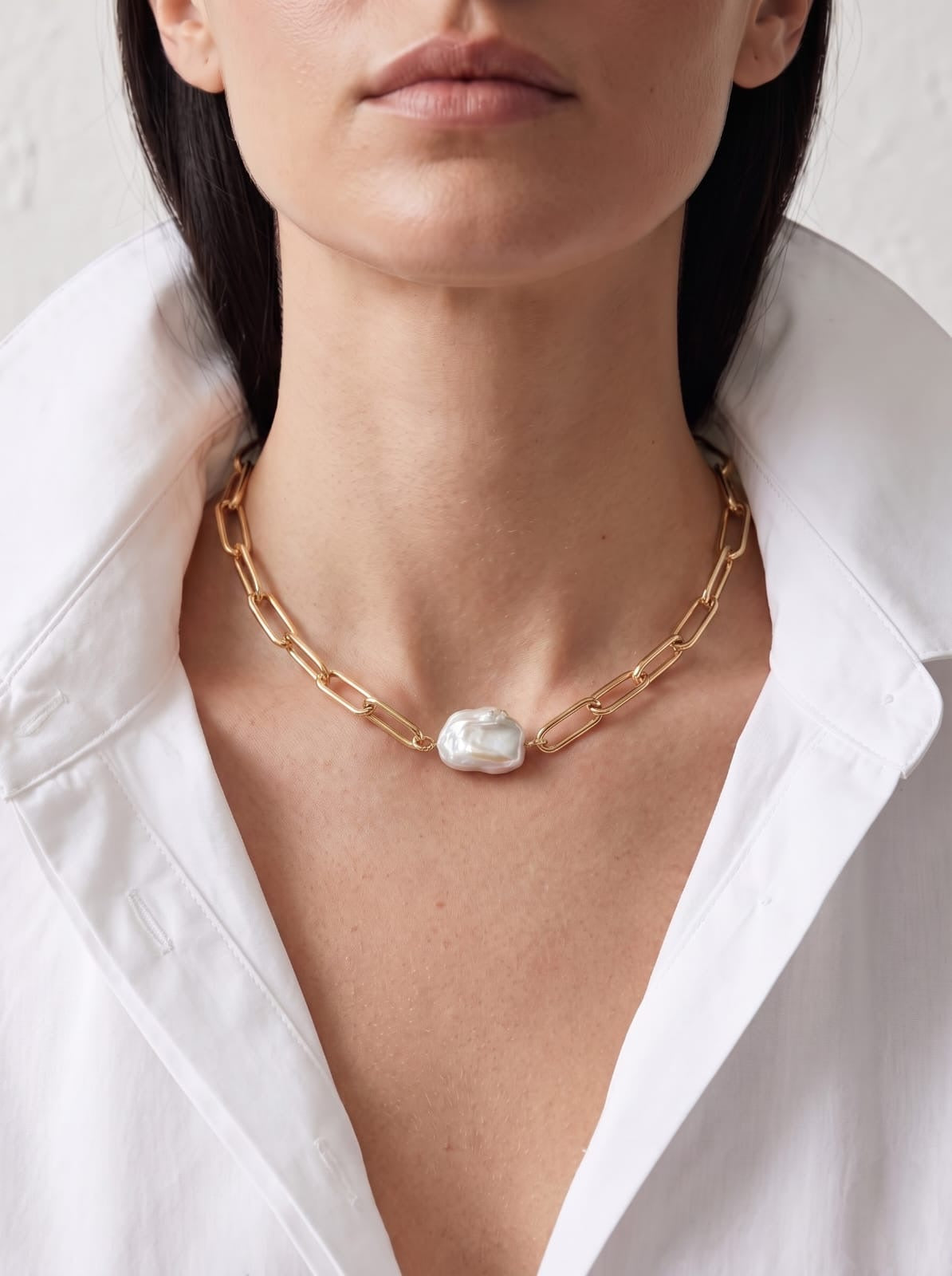 A close-up of a model wearing a gold paperclip chain necklace with a large, irregular white pearl pendant, styled with a white collared shirt.